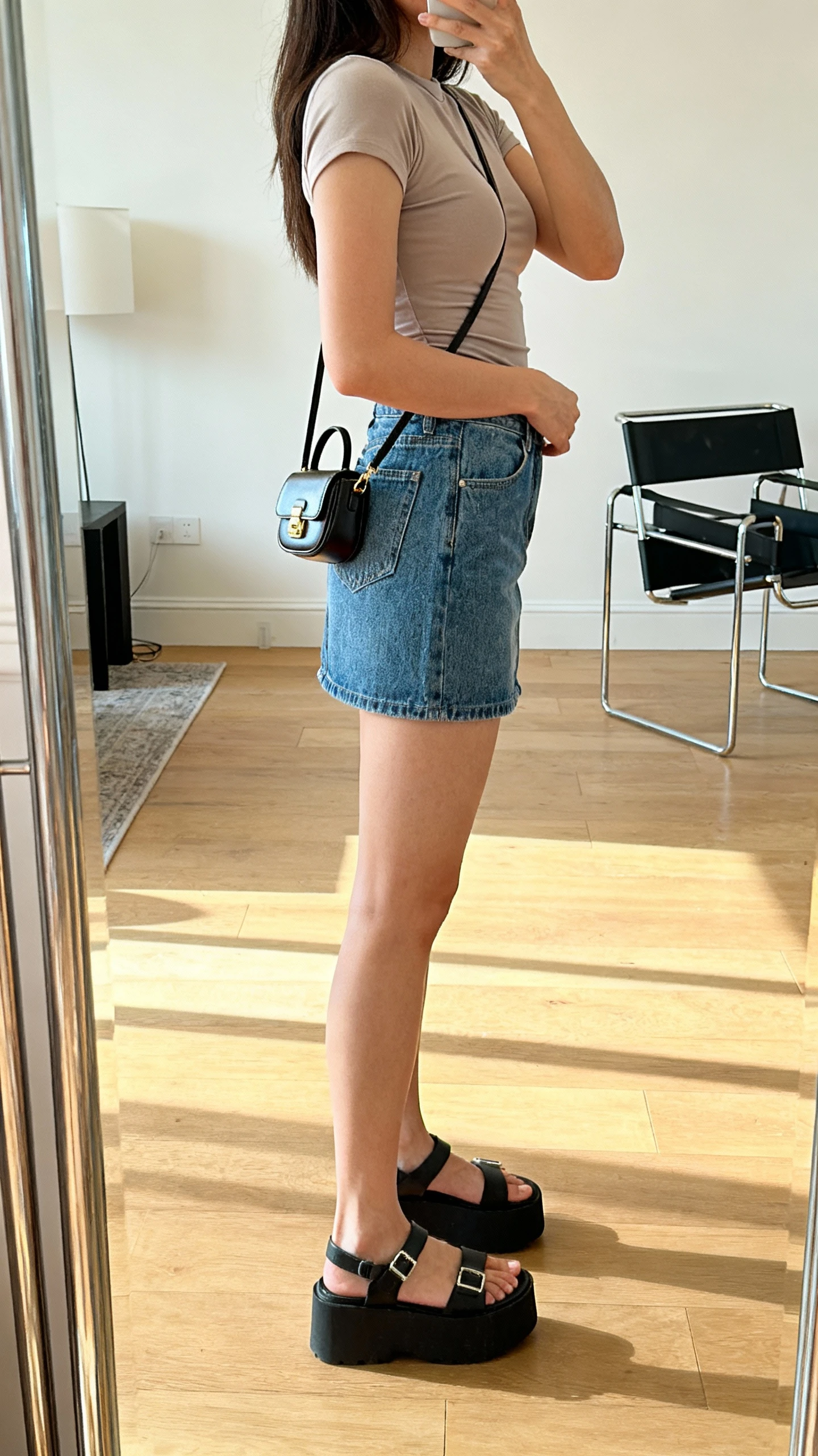 iPhone mirror selfie of a woman wearing a fitted baby tee, denim mini skirt, platform sandals, and a tiny micro bag on shoulder, face not visible, side angle, studio apartment mirror, bright daylight, iPhone photo quality.