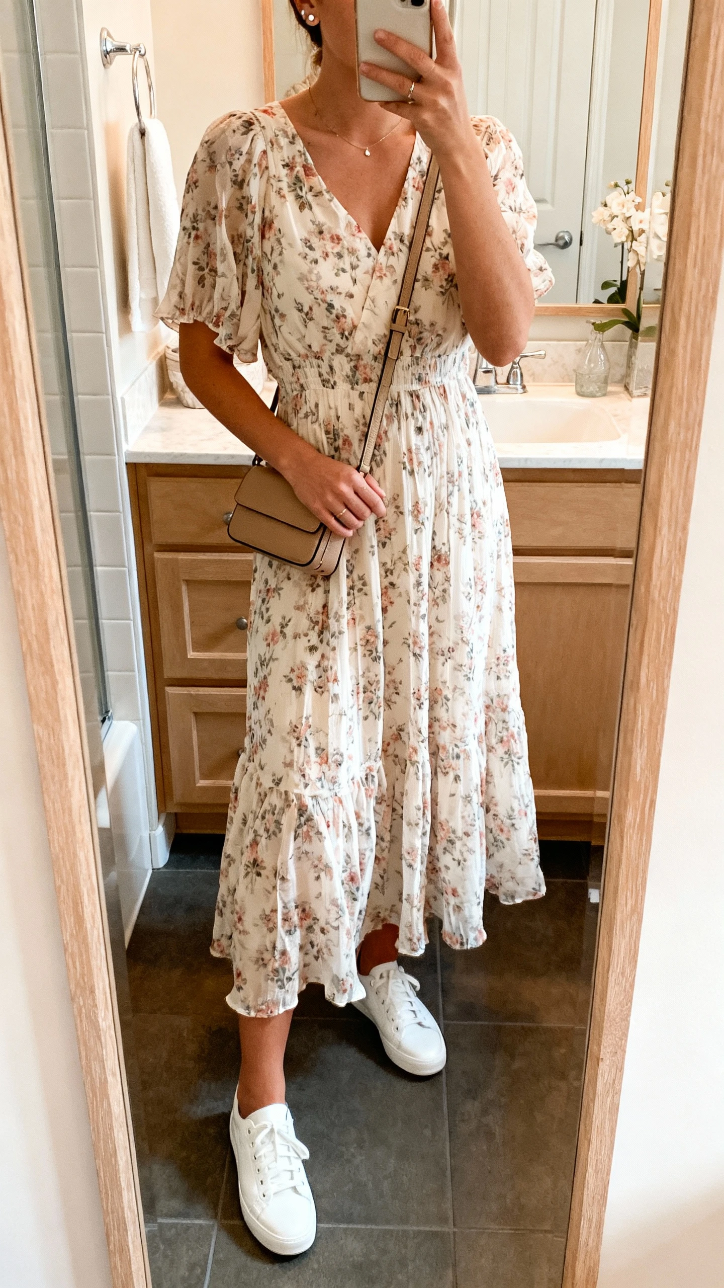 iPhone mirror selfie of a woman wearing a floaty floral midi dress with white low-top sneakers, crossbody bag, and delicate studs, face not visible, bathroom full-length mirror, soft morning light, iPhone photo quality.