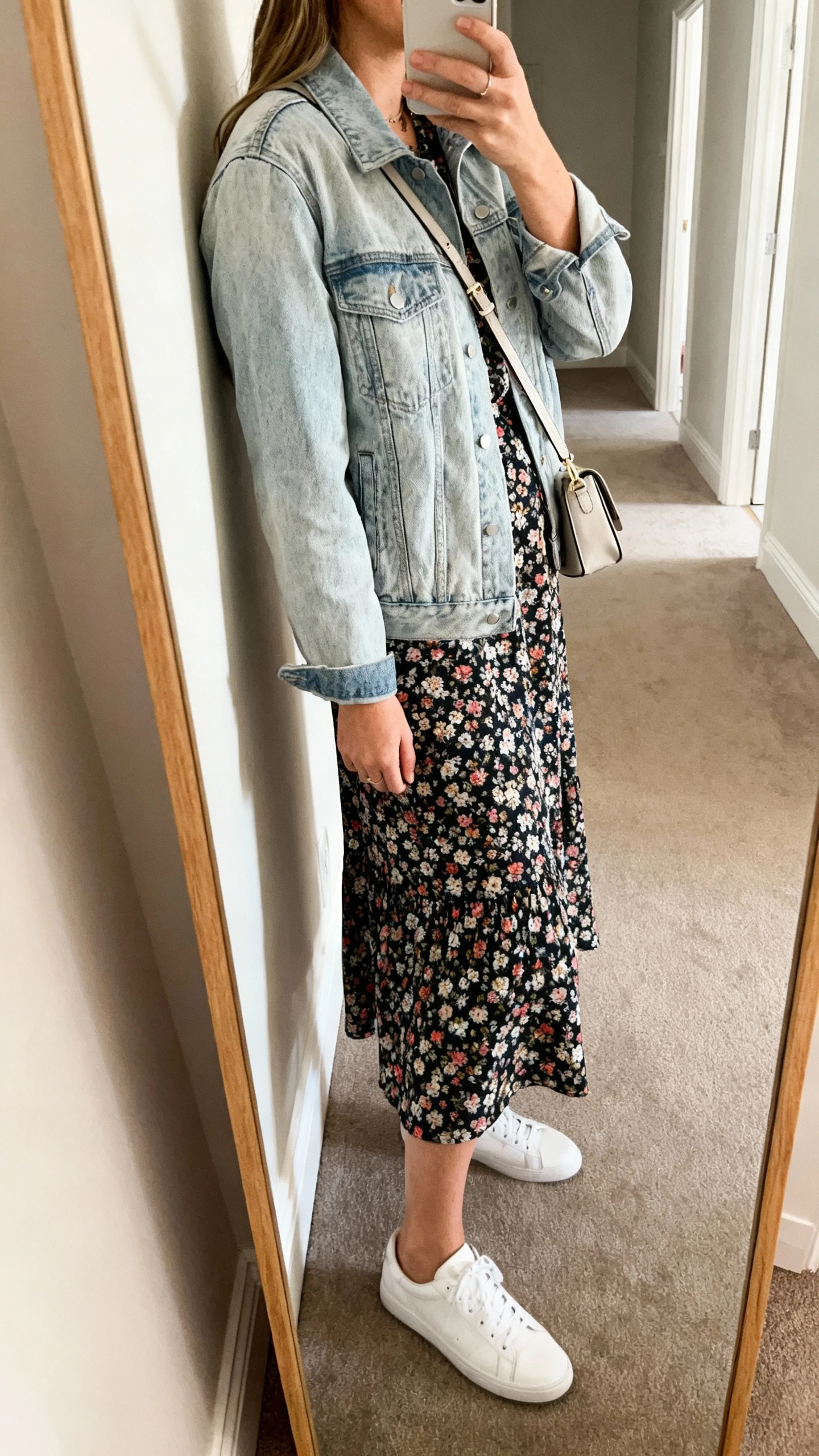 iPhone mirror selfie of a woman wearing a floral midi dress layered with a light-wash denim jacket, white sneakers, and a small crossbody bag, face not visible, side angle, full-length mirror in hallway, soft indoor daylight, iPhone photo quality.