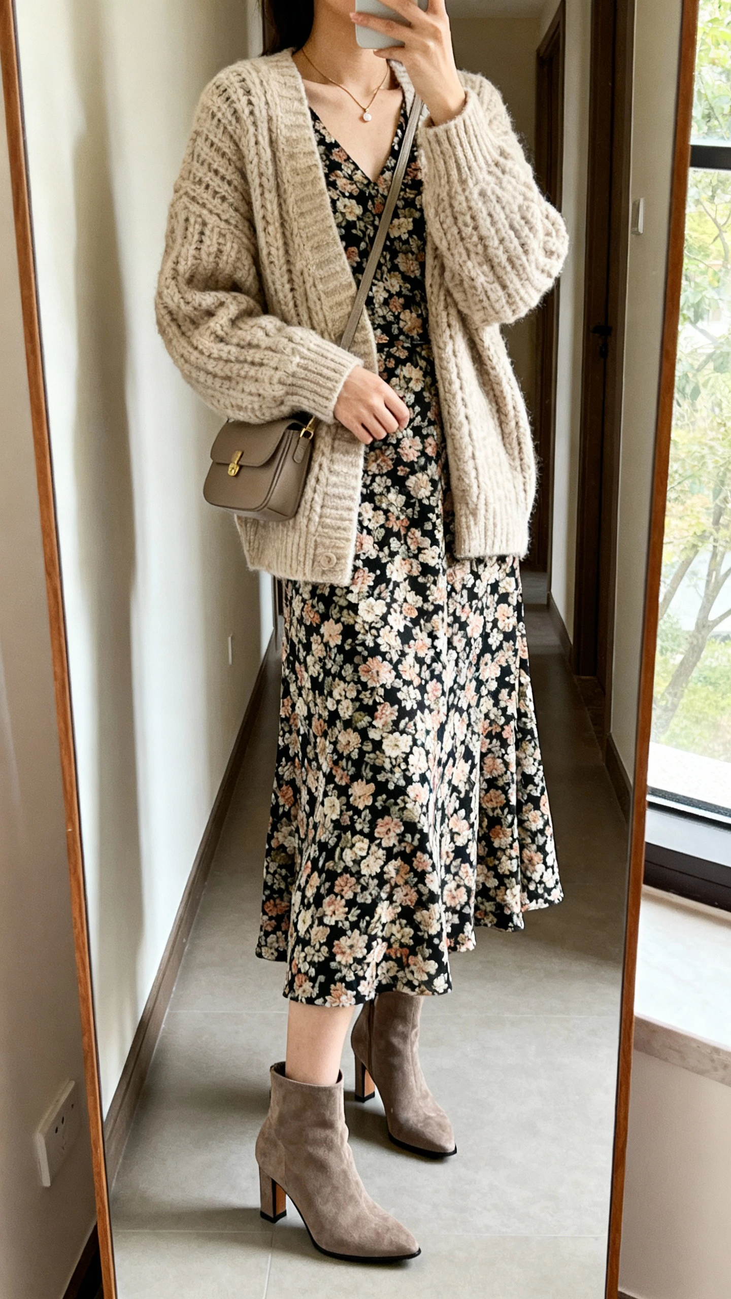 iPhone mirror selfie of a woman wearing a floral midi dress under a chunky oatmeal cardigan, mid-heel ankle boots, delicate necklace, and a small shoulder bag, face not visible, cropped at neck, full-length mirror in hallway, soft window light, iPhone photo quality.