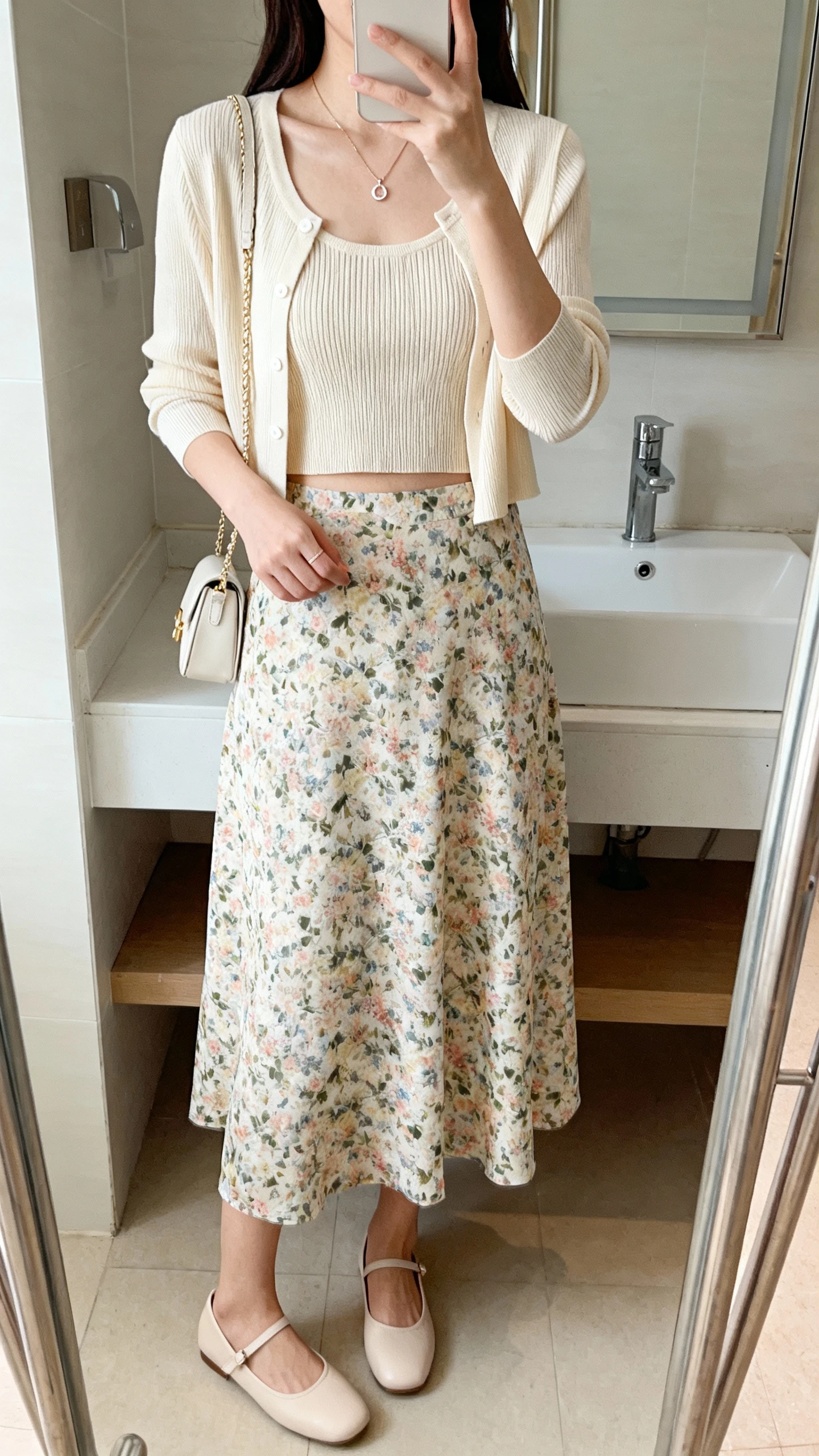 iPhone mirror selfie of a woman wearing a floral midi skirt with a cropped cardigan set (cardigan plus matching knit tank), ballet flats, dainty pendant, small shoulder bag, face not visible, bathroom mirror, natural daylight, iPhone photo quality.