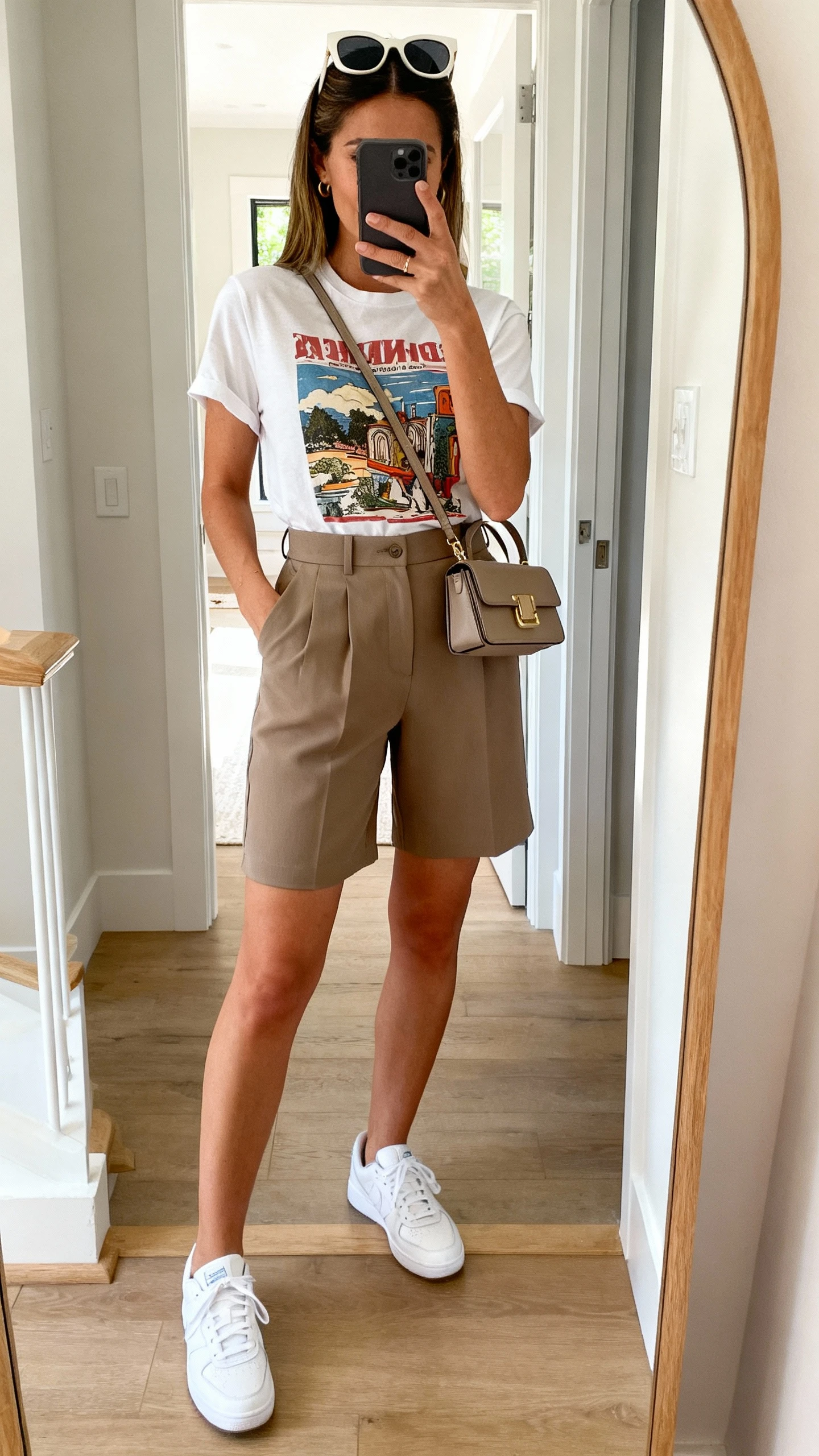 iPhone mirror selfie of a woman wearing a graphic tee tucked into tailored high-waist shorts, clean white sneakers, oversized sunglasses on head, structured mini bag, face not visible, cropped at shoulders, entryway mirror, natural daylight, iPhone photo quality.