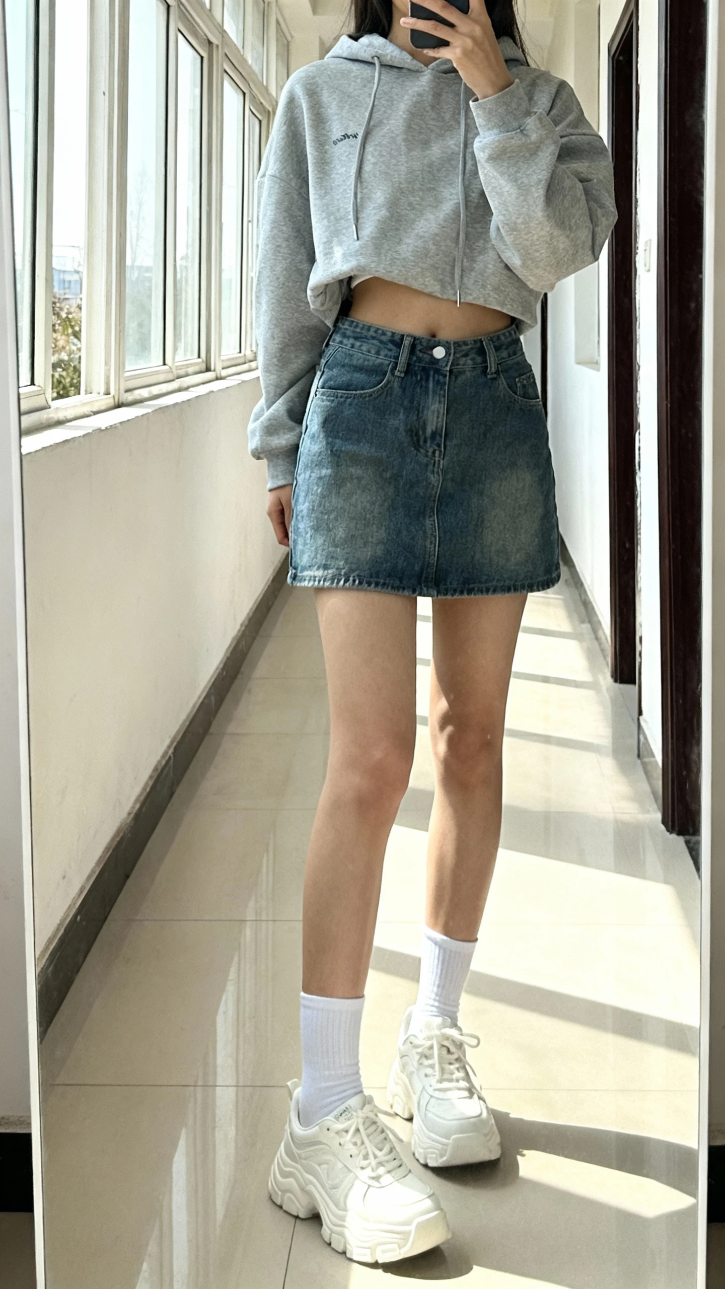 iPhone mirror selfie of a woman wearing a gray hoodie half-tucked into a denim mini skirt with white crew socks and chunky white sneakers, face not visible, cropped at shoulders, full-length mirror in hallway, bright daylight from window, iPhone photo quality.