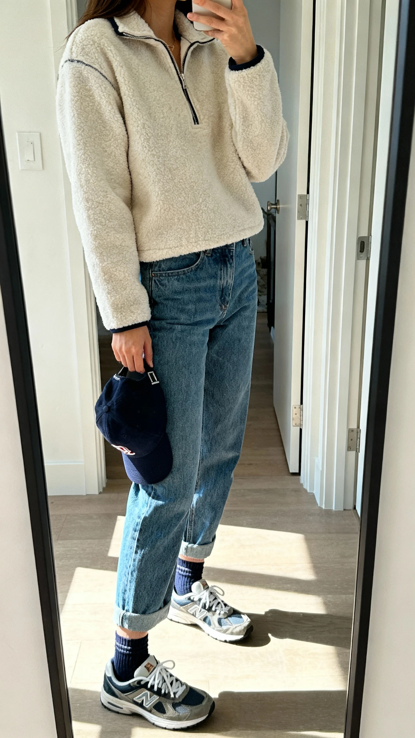iPhone mirror selfie of a woman wearing a half-zip fleece pullover with mom jeans, retro sneakers, ankle socks, and a baseball cap in hand, face not visible, side angle, entryway mirror, bright natural light, iPhone photo quality.