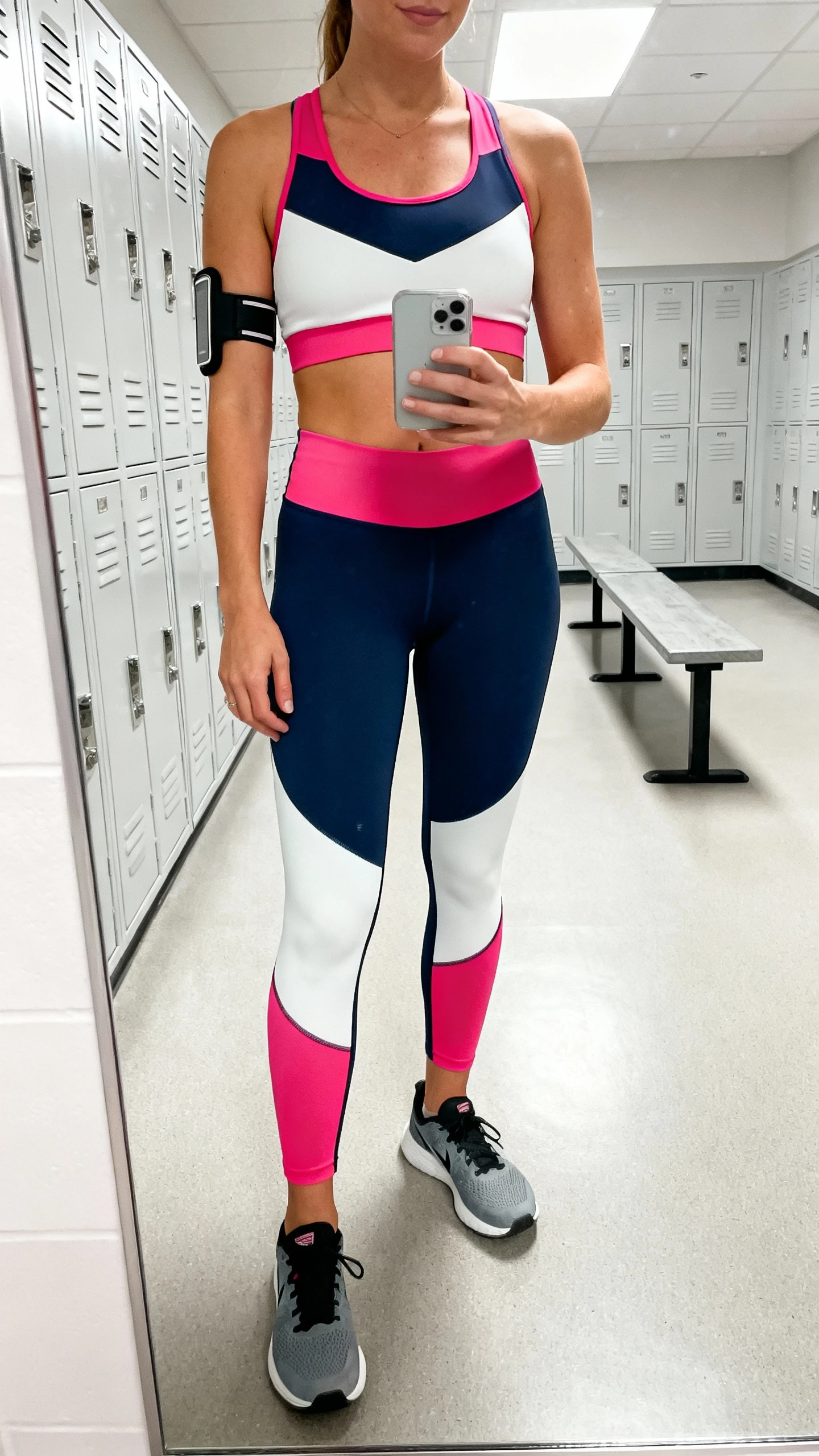 iPhone mirror selfie of a woman wearing a high-performance mix with strategic color blocking: color-block compression bra, matching color-block leggings, stability running shoes, and a slim arm band for phone, face not visible, cropped at shoulders, gym locker-room mirror, bright overhead light, iPhone photo quality