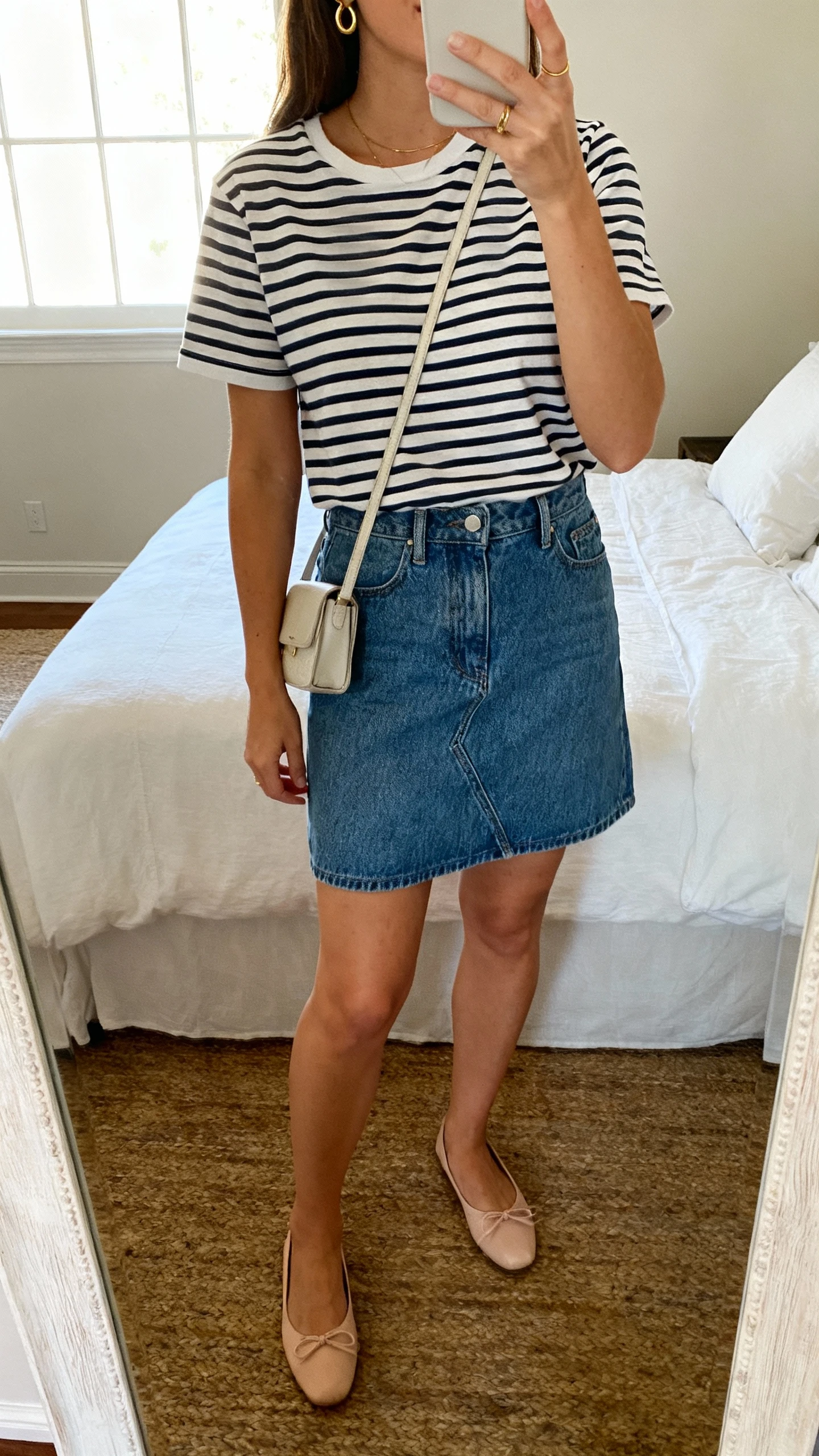 iPhone mirror selfie of a woman wearing a jeans mini skirt with a French-girl striped tee and ballet flats, small crossbody bag, delicate gold hoops, face not visible, cropped at neck, bedroom mirror, natural window light, iPhone photo quality.