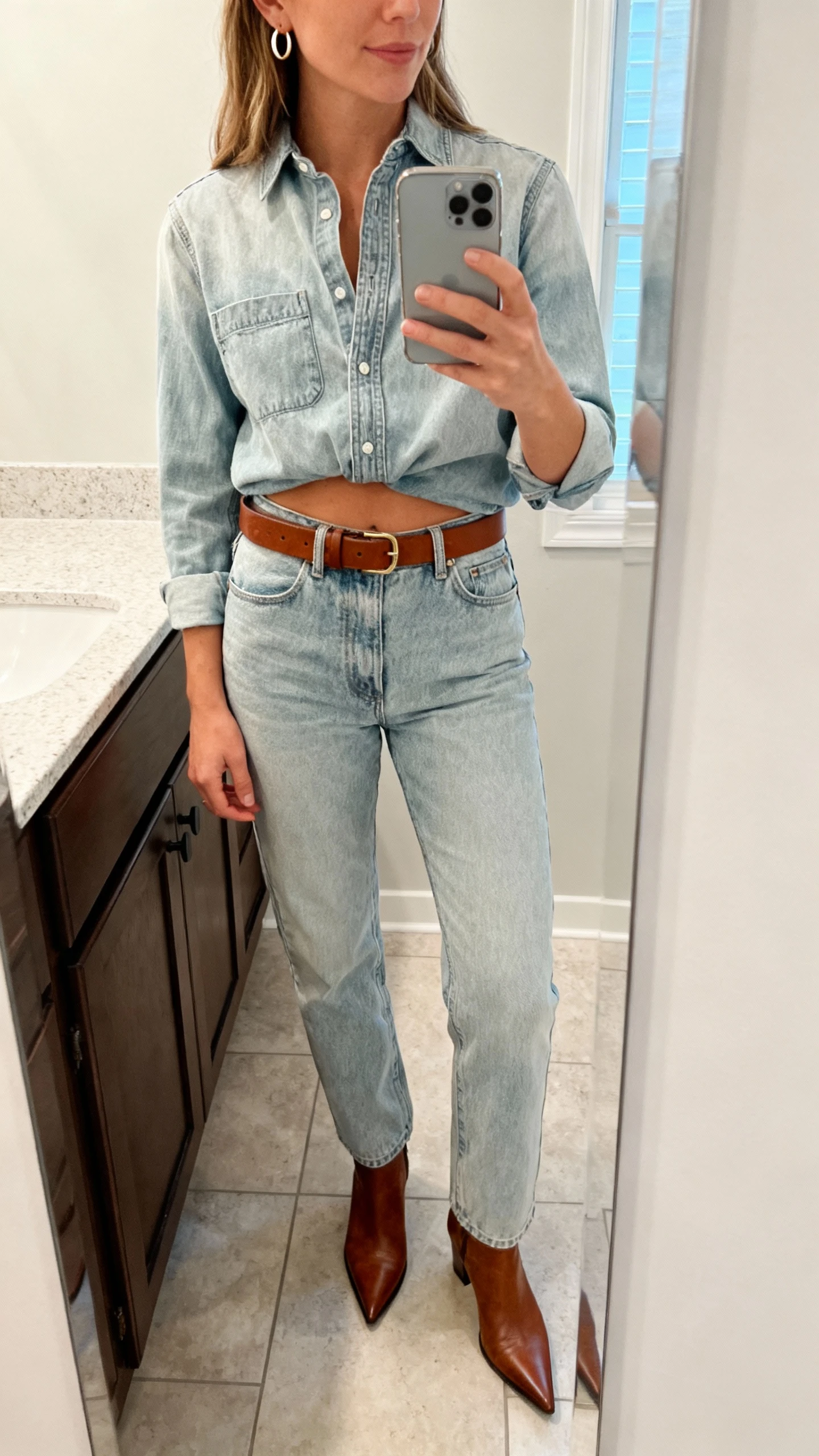 iPhone mirror selfie of a woman wearing a light-wash denim shirt half-tucked into matching light-wash straight jeans, brown leather belt, pointed ankle boots, small hoop earrings, face not visible, bathroom mirror, natural indoor light, iPhone photo quality