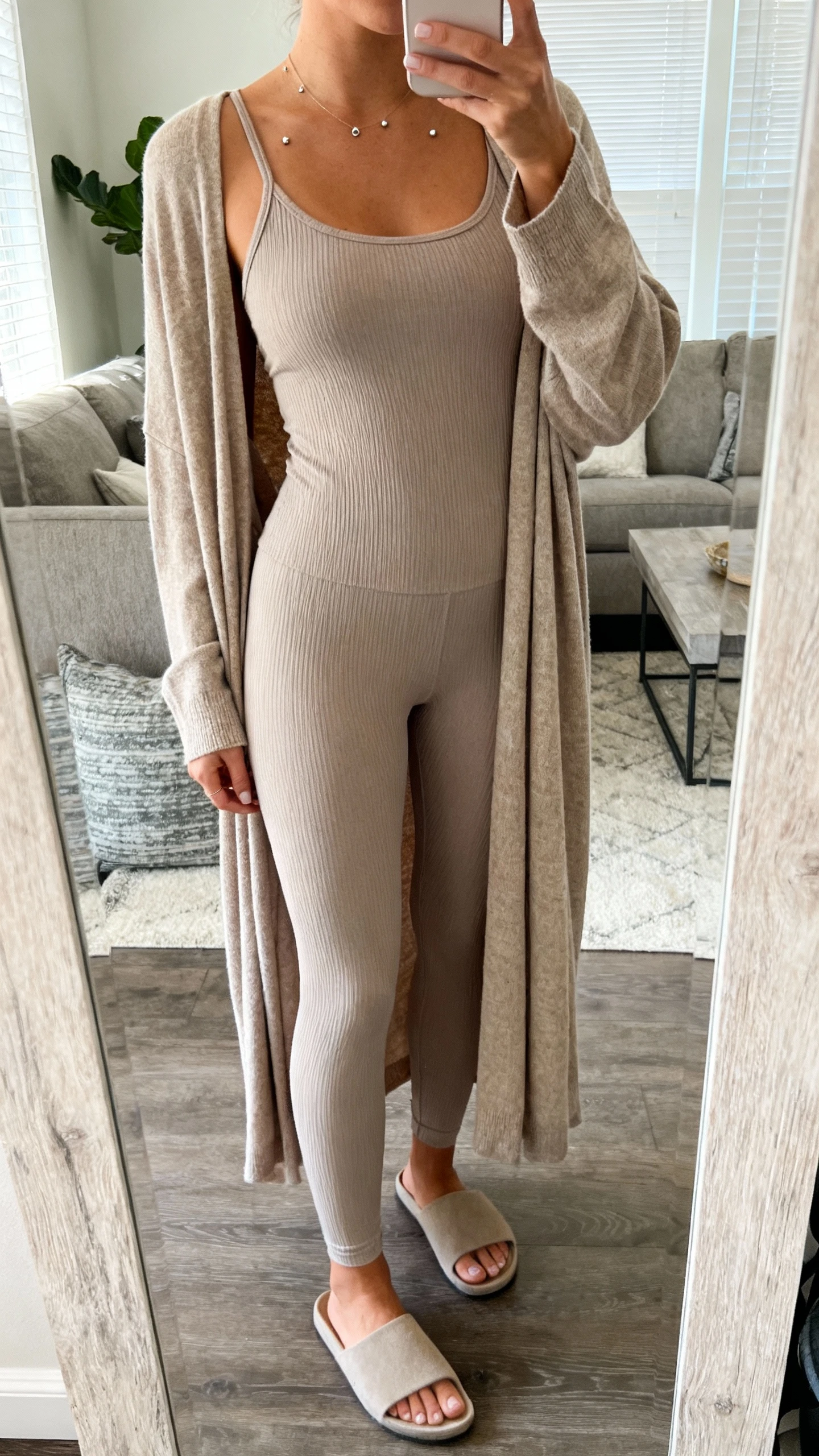 iPhone mirror selfie of a woman wearing a long, drapey cardigan over a fitted matching knit tank and leggings for a column look, delicate studs, minimalist slides, face not visible, cropped at shoulders, living room mirror, natural morning light, iPhone photo quality.