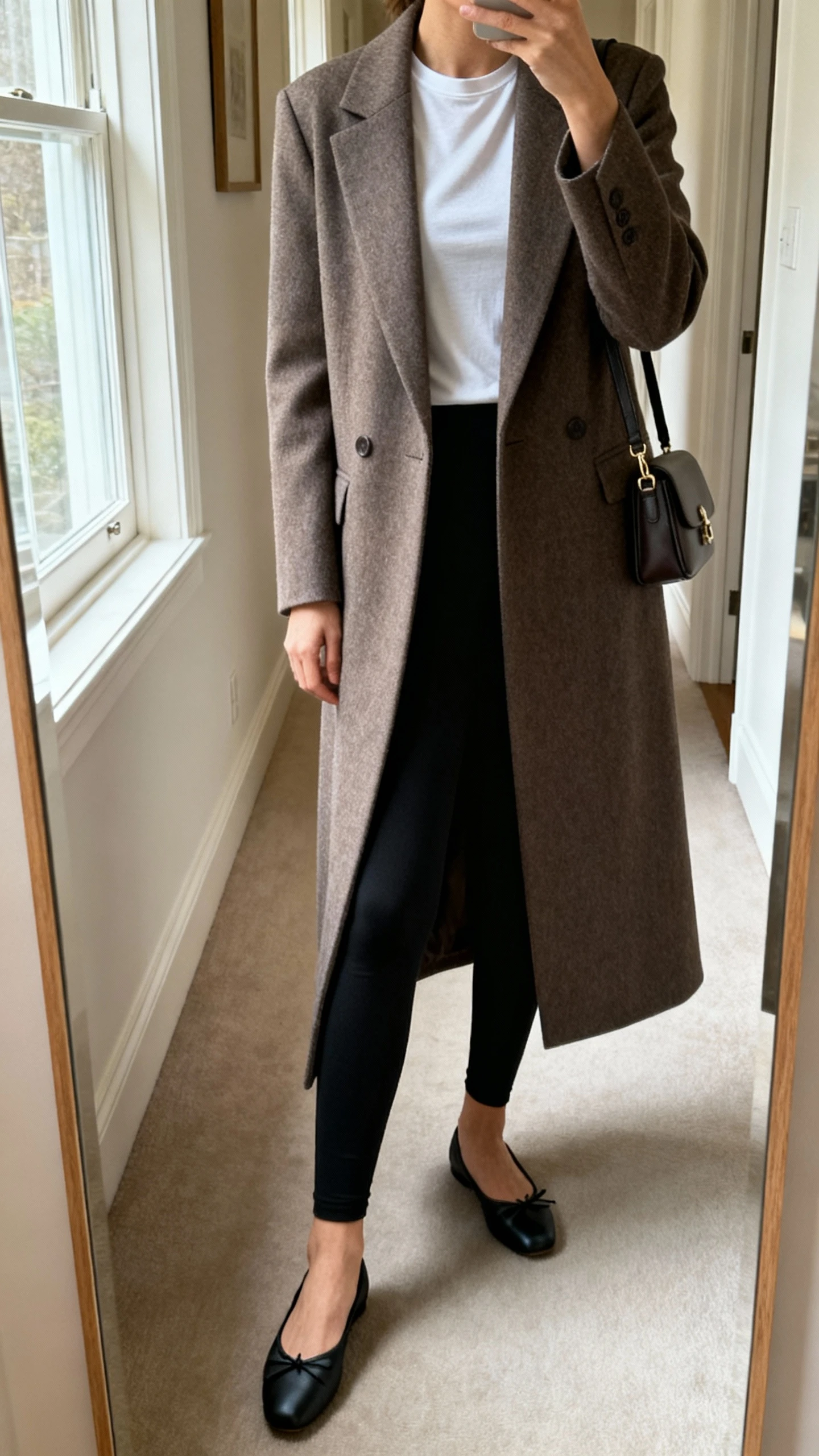 iPhone mirror selfie of a woman wearing a long tailored coat over simple black leggings and a fitted tee, classic ballet flats, small shoulder bag, face not visible, cropped at neck, hallway full-length mirror, soft morning window light, iPhone photo quality.