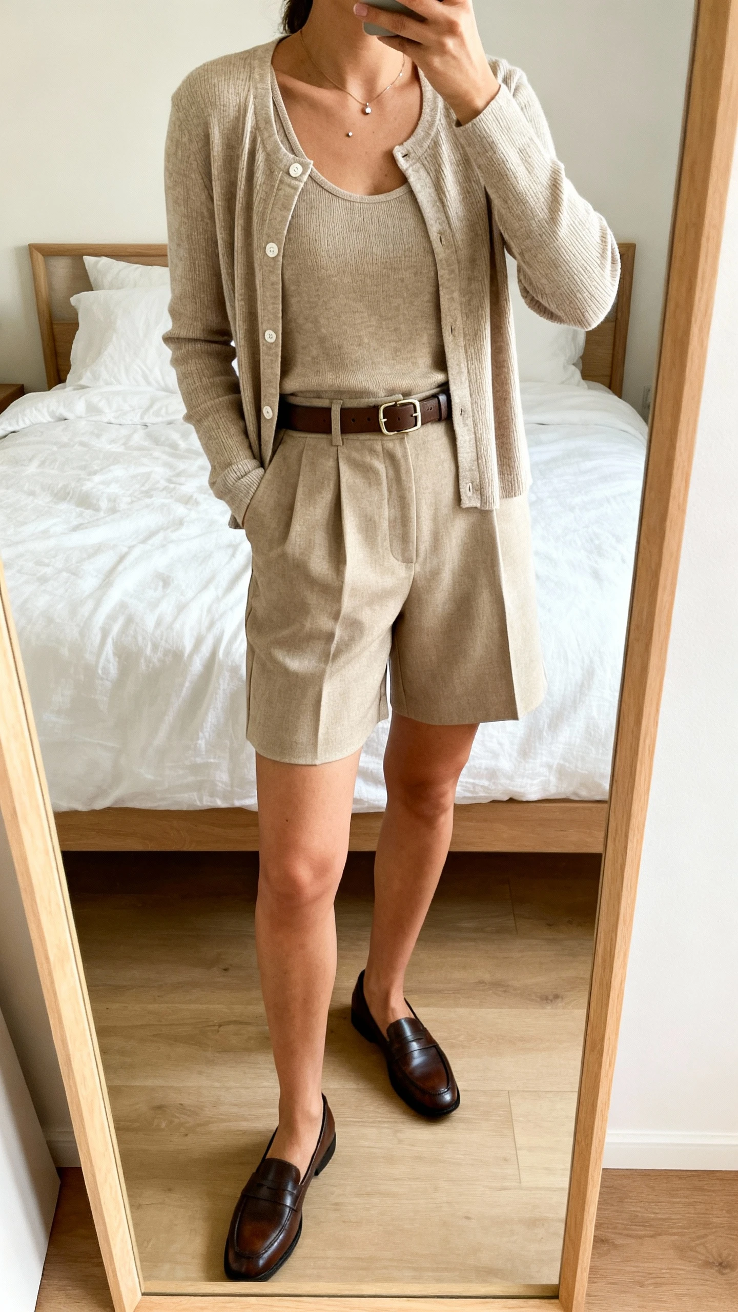 iPhone mirror selfie of a woman wearing a matching cardigan set (tank + cardigan) tucked into tailored high-waisted shorts, loafers, belt, and simple studs, face not visible, cropped at shoulders, full-length mirror in bedroom, soft daylight, iPhone photo quality.