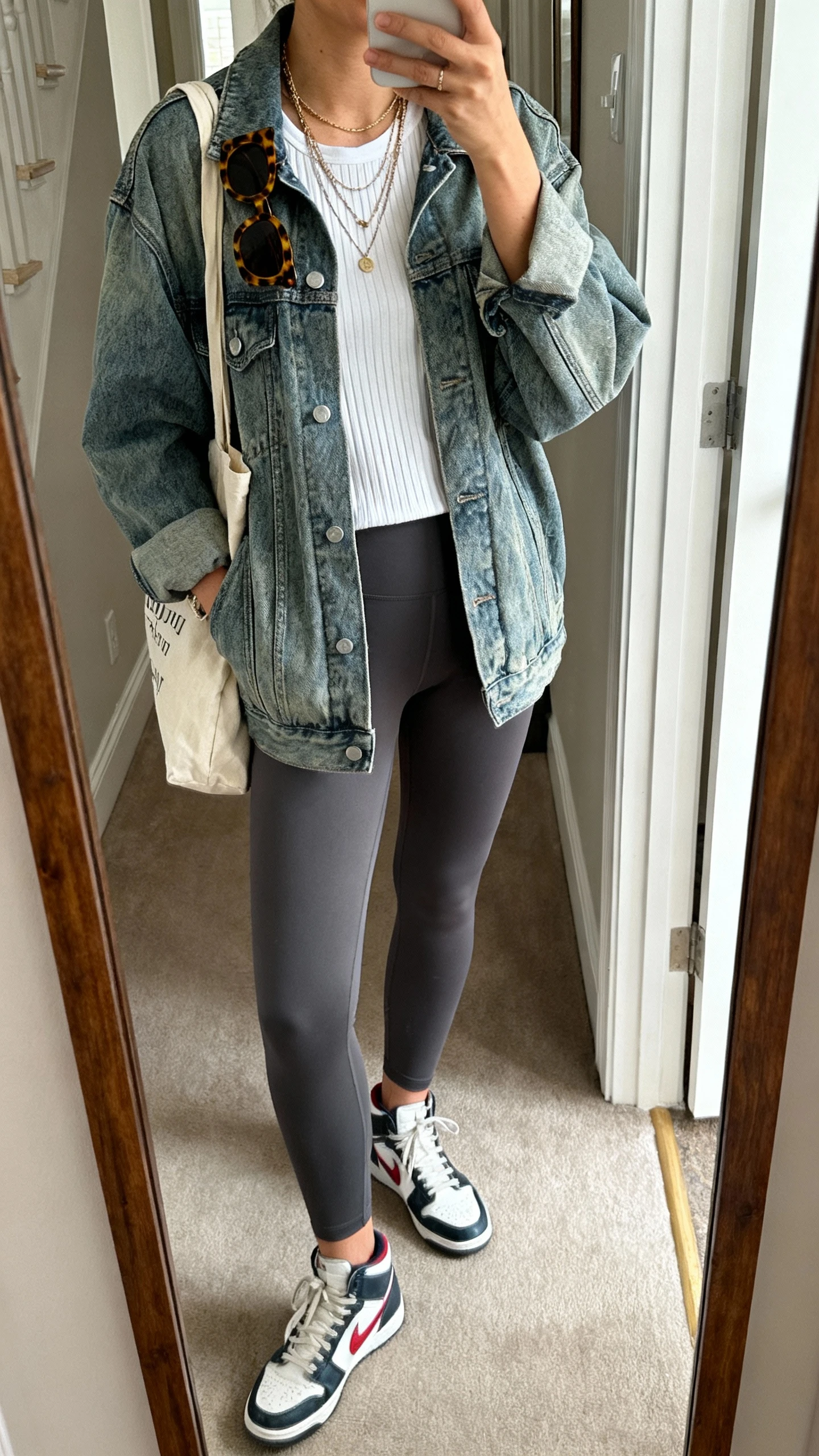 iPhone mirror selfie of a woman wearing a mid-wash slightly oversized denim jacket, charcoal leggings, white ribbed tee, retro court sneakers, tortoiseshell sunglasses tucked on collar, canvas tote bag, layered necklaces, face not visible, cropped at shoulders, entryway full-length mirror, natural daylight, iPhone photo quality.