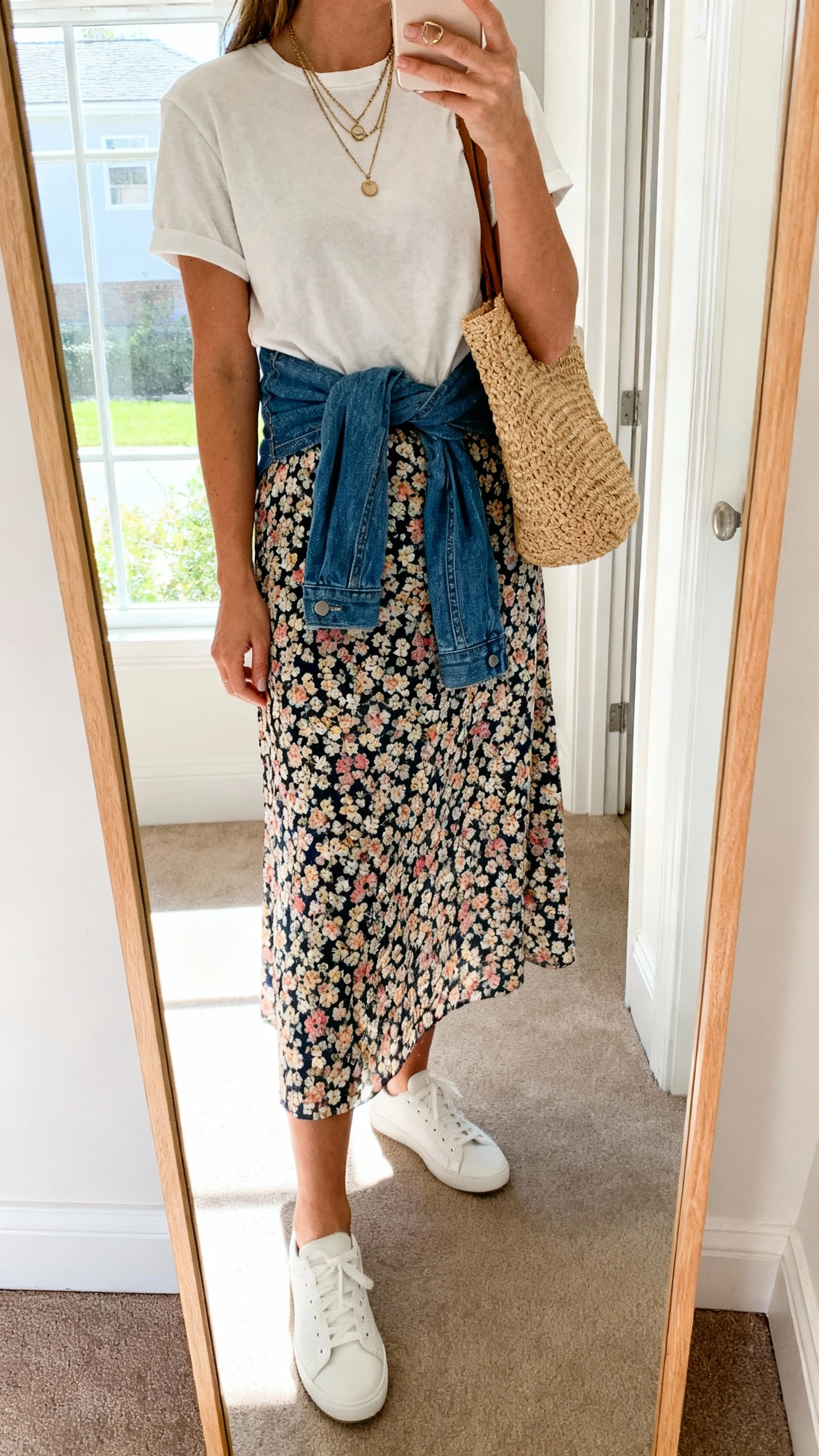 iPhone mirror selfie of a woman wearing a midi floral skirt with a tucked-in relaxed cotton tee, denim jacket tied around the waist, white low-top sneakers, straw tote, and layered necklaces, face not visible, cropped at neck, entryway mirror, bright window light, iPhone photo quality