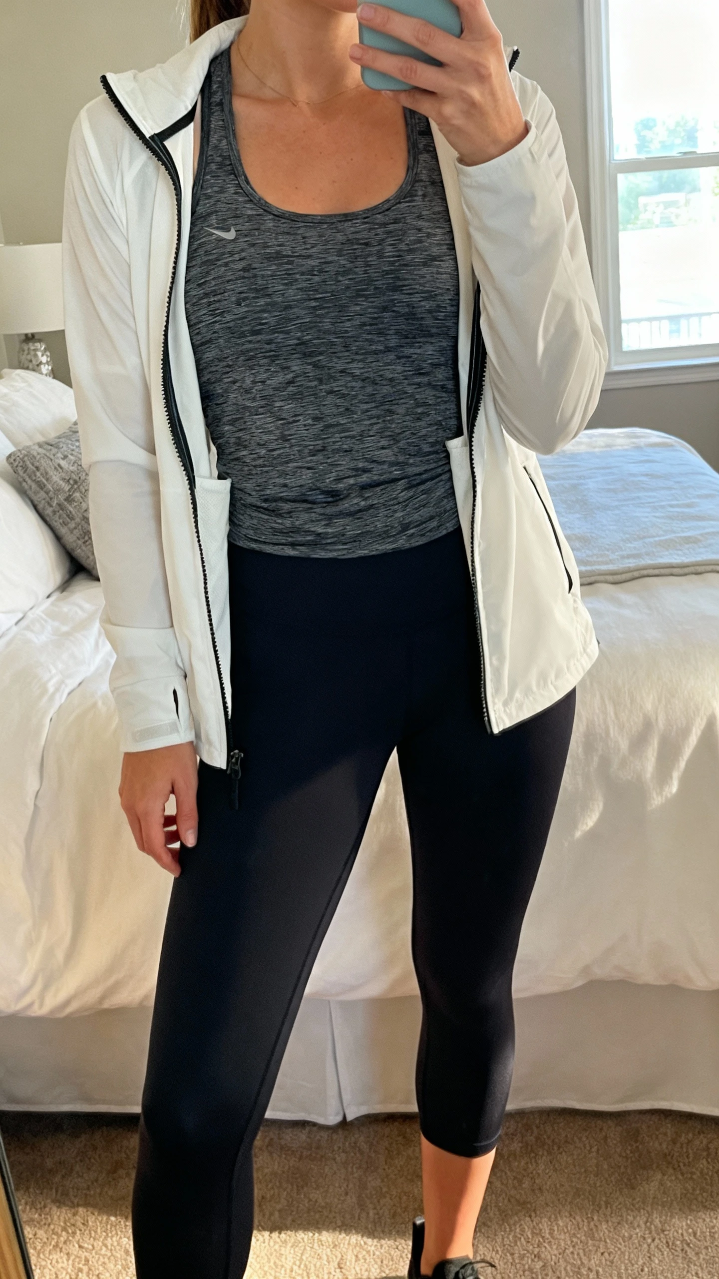 iPhone mirror selfie of a woman wearing a moisture-wicking base tank, lightweight zip-up running jacket, and 7/8 leggings, with the jacket unzipped for layering, face not visible, cropped at neck, bedroom mirror, morning window light, iPhone photo quality