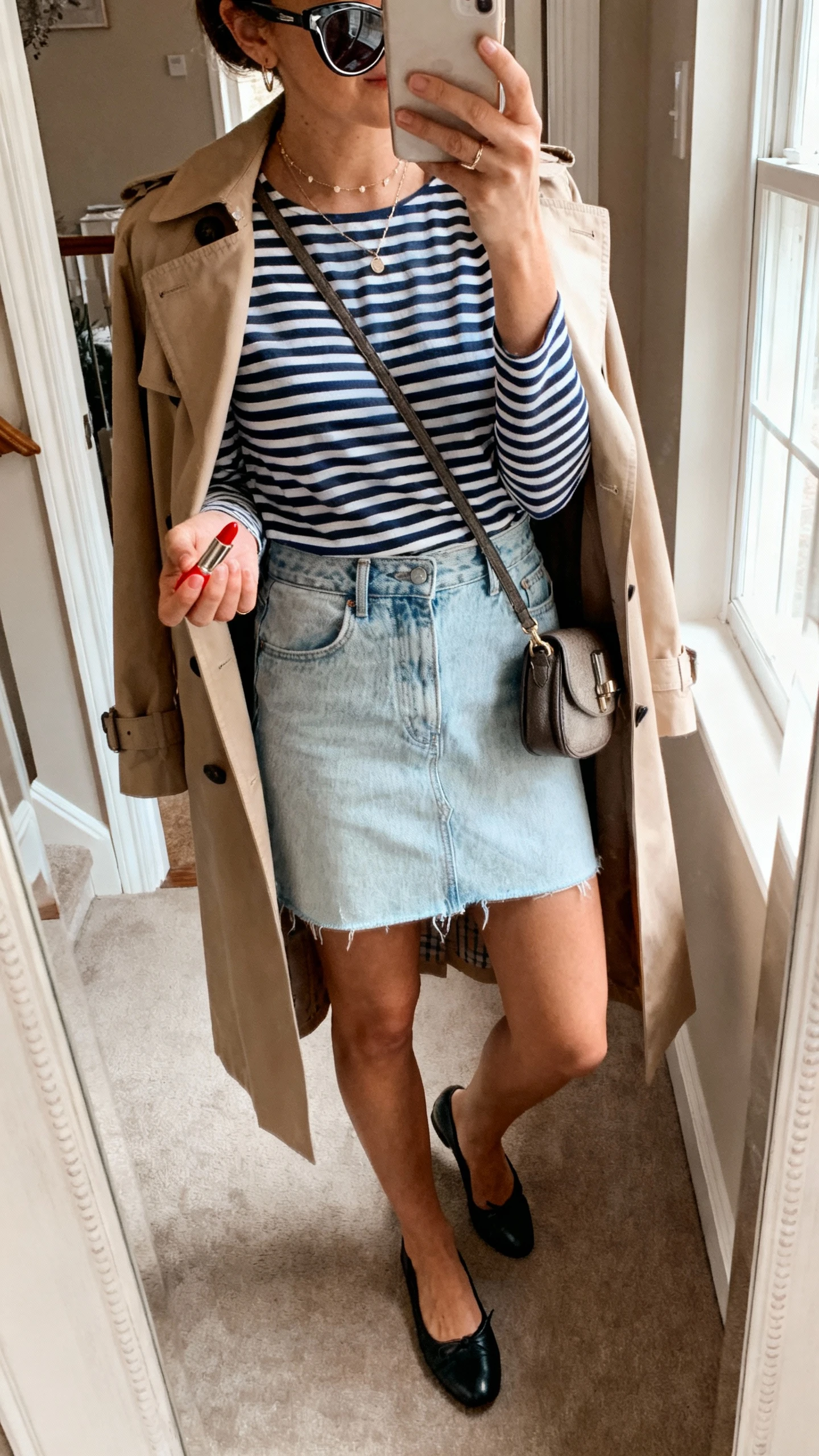 iPhone mirror selfie of a woman wearing a navy-and-white Breton striped top, light-wash denim mini skirt, black leather ballet flats, classic trench coat, small shoulder bag, red lipstick in hand and cat-eye sunglasses on head, dainty jewelry, face not visible, cropped at neck, entryway mirror, indoor window light, iPhone photo quality.
