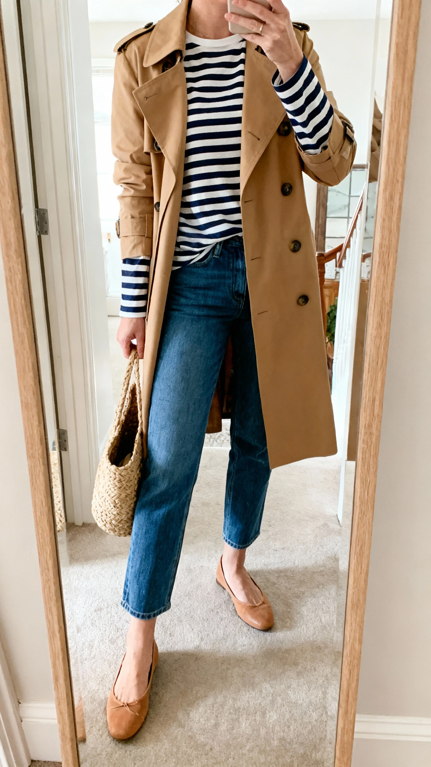 iPhone mirror selfie of a woman wearing a navy-and-white Breton striped long-sleeve tee under a camel trench coat with cropped straight blue jeans, tan ballet flats, woven tote, face not visible, cropped at neck, entryway mirror, soft morning light, iPhone photo quality