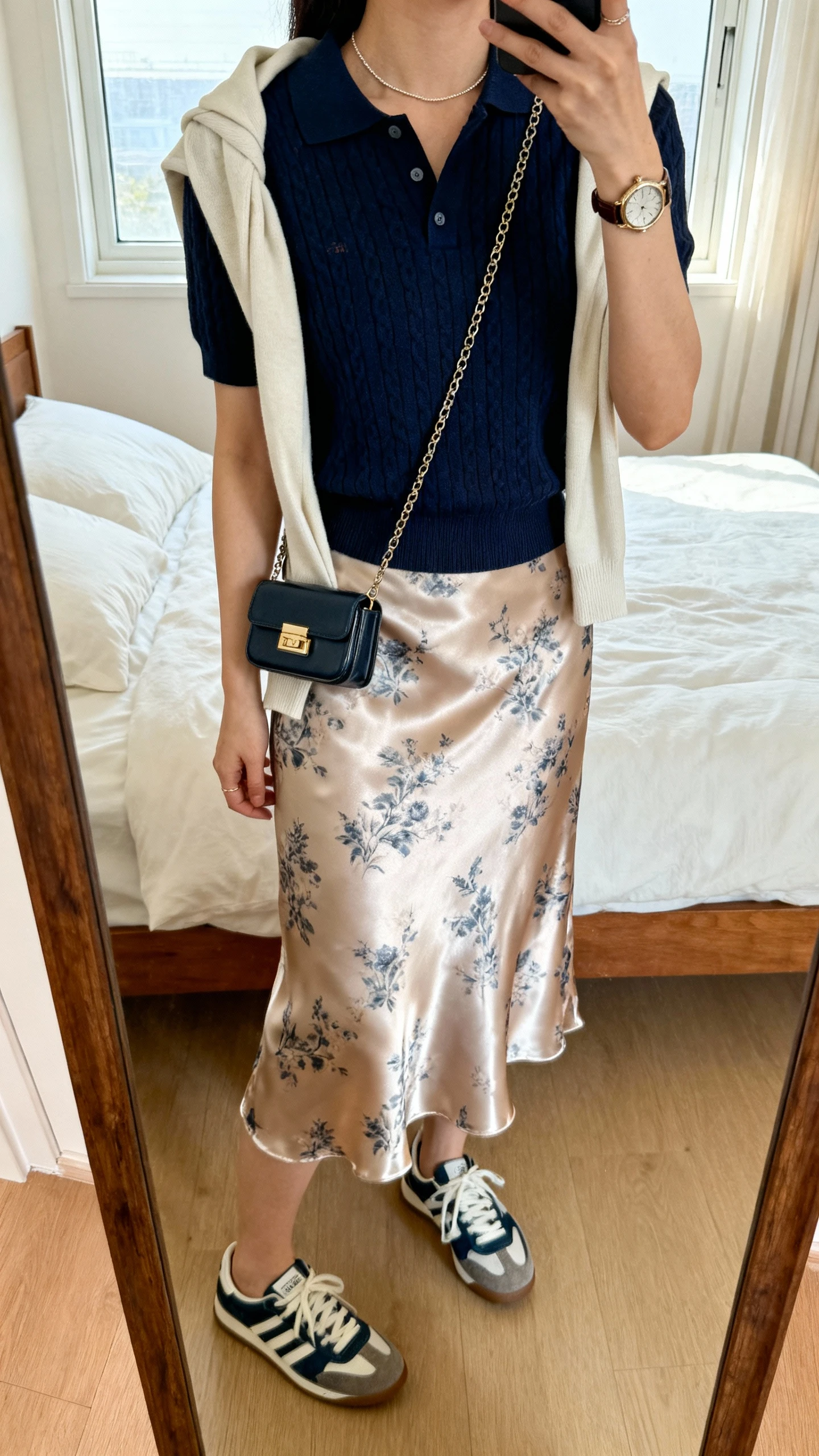 iPhone mirror selfie of a woman wearing a navy short-sleeve knit polo, satin midi slip skirt (printed), retro sneakers, compact shoulder bag, thin chain necklace, vintage-inspired watch, light cardigan tied around shoulders; face not visible, cropped at neck, bedroom mirror, natural window light, iPhone photo quality.