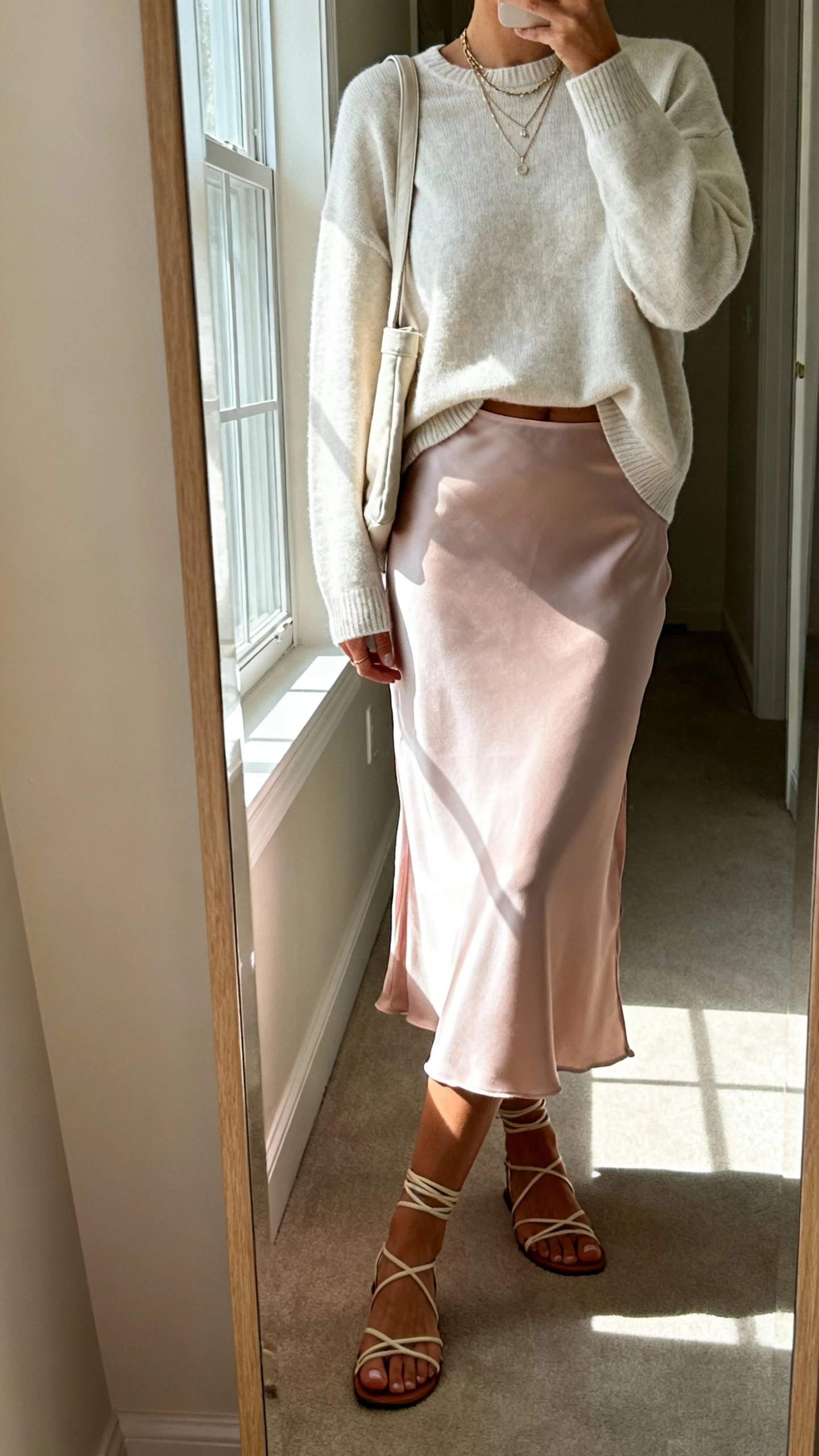 iPhone mirror selfie of a woman wearing a pastel satin midi slip skirt, lightweight neutral crewneck sweater half-tucked, strappy sandals, delicate layered necklaces, and a soft shoulder bag; face not visible, cropped at shoulders, full-length mirror in hallway with window light, iPhone photo quality.
