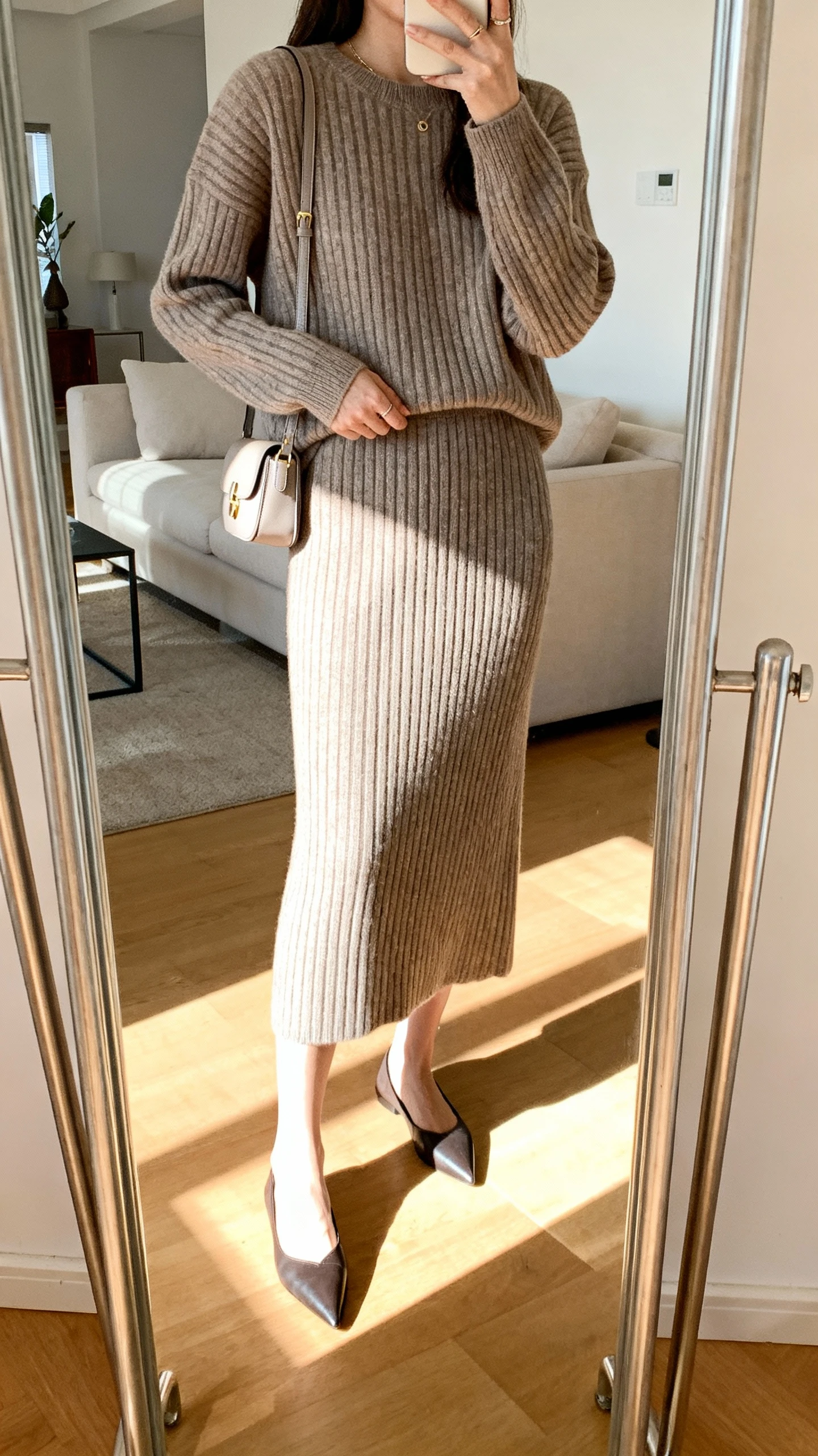 iPhone mirror selfie of a woman wearing a polished knit set (matching ribbed sweater and midi skirt) with pointed flats and a small shoulder bag, face not visible, cropped at shoulders, living room mirror, bright indoor light, iPhone photo quality.