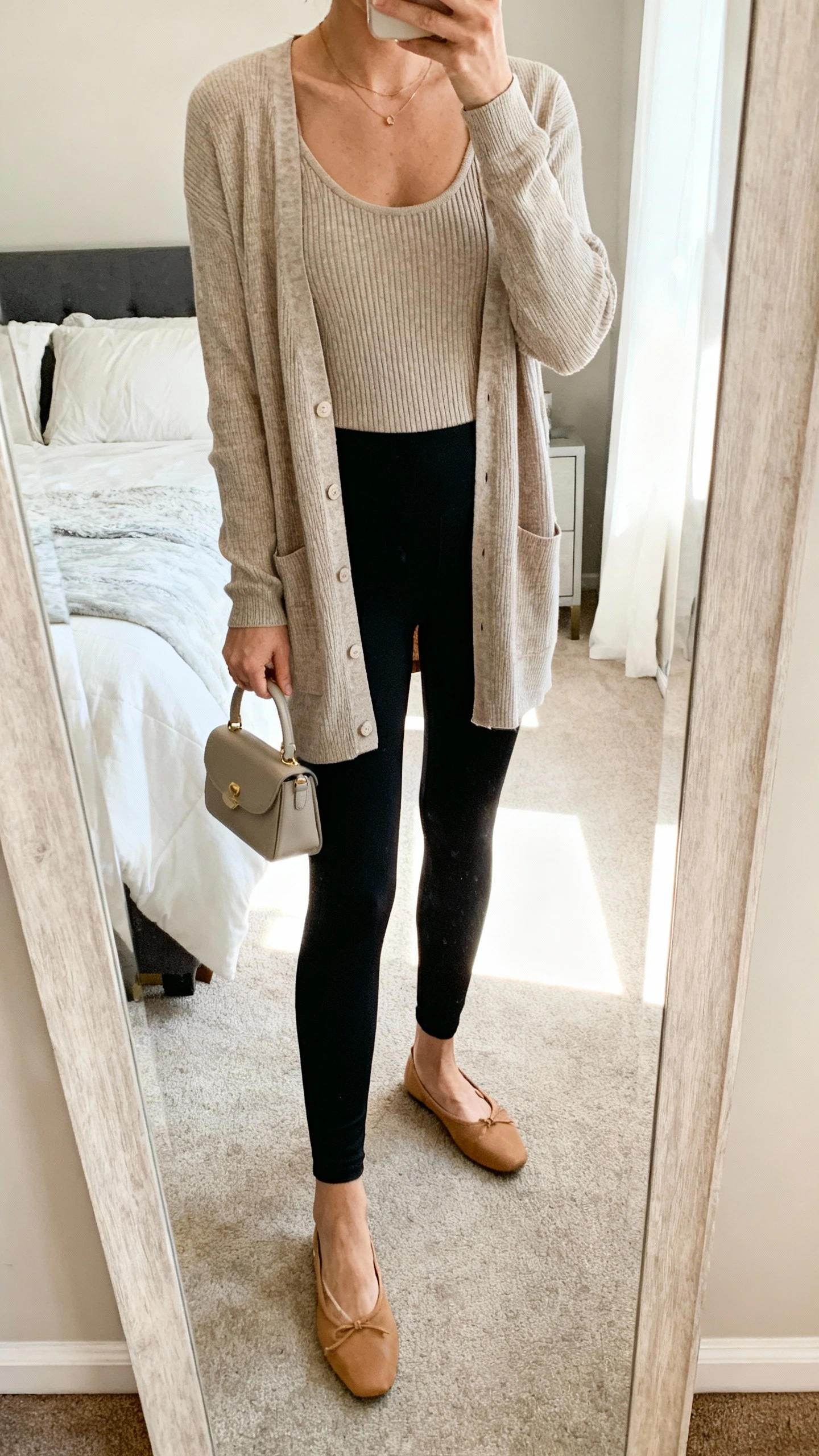 iPhone mirror selfie of a woman wearing a polished cardigan twinset (fine-knit tank with matching cardigan), ankle-length black leggings, tan ballet flats, and a small top-handle bag, face not visible, cropped at neck, bedroom full-length mirror, soft morning light, iPhone photo quality.