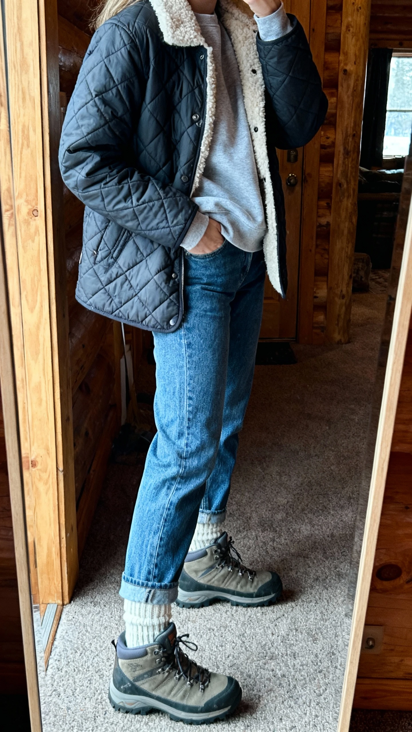 iPhone mirror selfie of a woman wearing a quilted jacket layered over a sherpa-lined sweatshirt with mom jeans, wool socks, and hiking-inspired boots, face not visible, side angle, cabin-style entry mirror, cool morning light, iPhone photo quality.