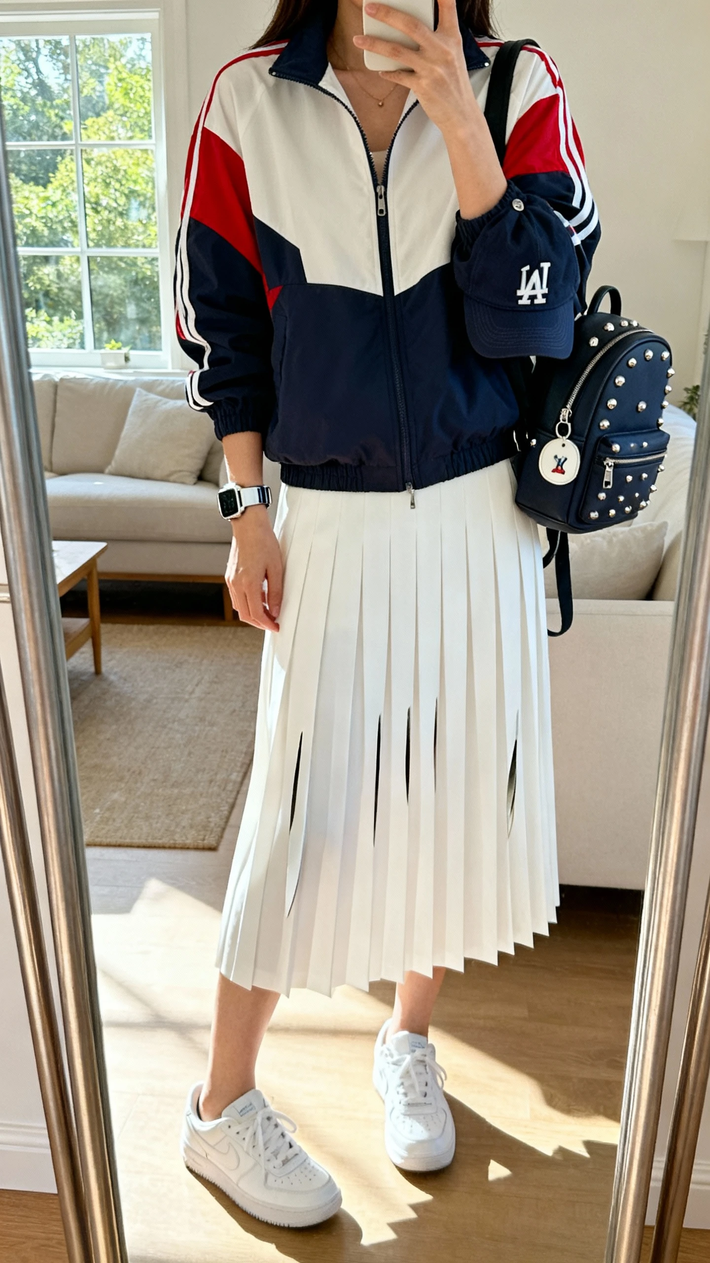 iPhone mirror selfie of a woman wearing a retro color-block track jacket, white knife-pleat midi skirt, sleek white trainers, compact backpack, baseball cap clipped to the bag, silver studs, and a sporty watch, face not visible, full-length mirror in a bright living room, natural window light, iPhone photo quality.
