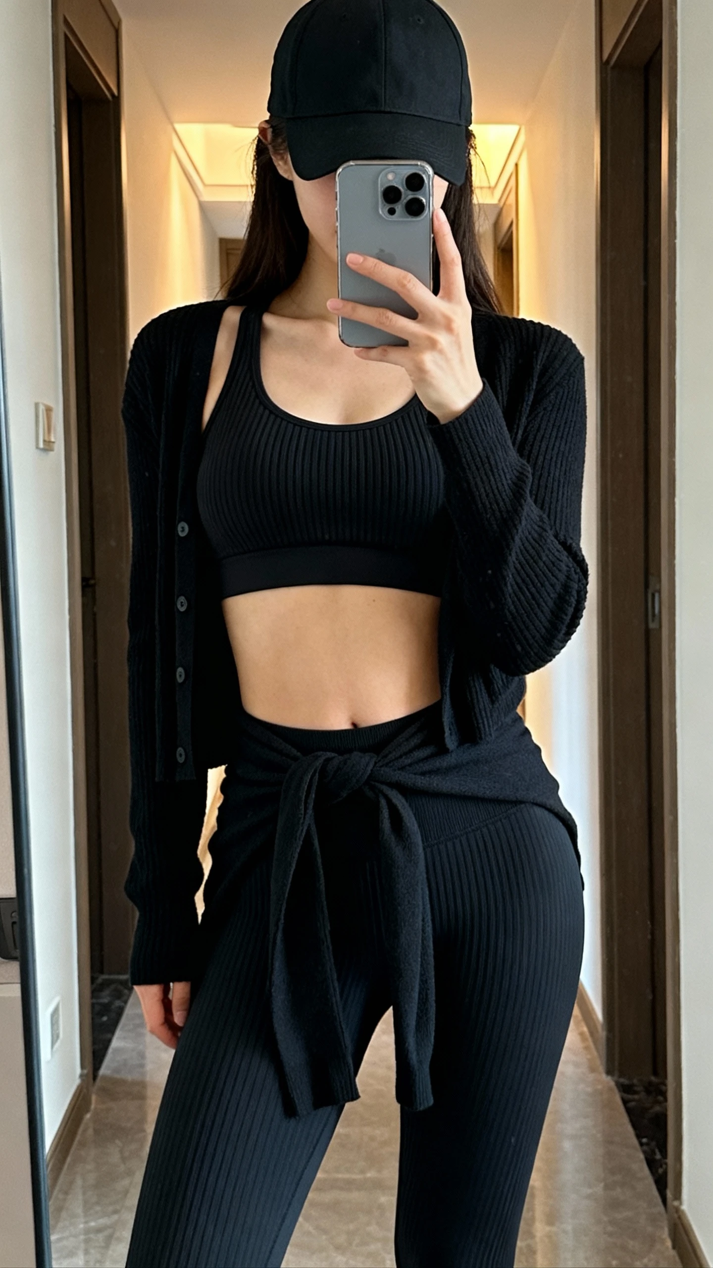 iPhone mirror selfie of a woman wearing a ribbed black workout set (ribbed bra and ribbed leggings) with tonal black cardigan tied at waist and a matte black cap, face not visible, cropped at shoulders, full-length mirror in hallway with indoor light, iPhone photo quality.