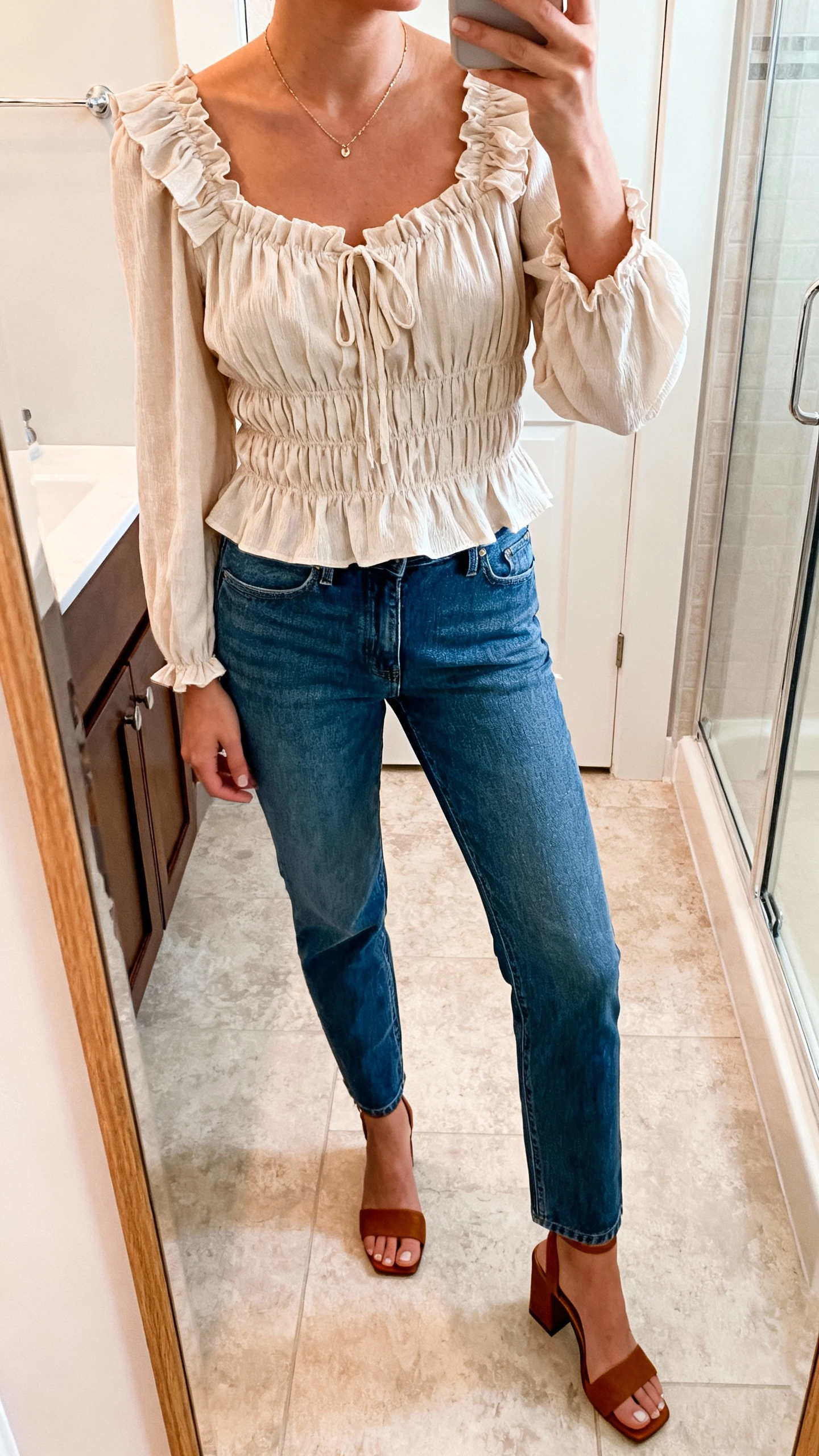 iPhone mirror selfie of a woman wearing a romantic blouse with ruffles/smocking tucked into straight-leg jeans, delicate necklace, and block-heel sandals, face not visible, cropped at neck, bathroom mirror, soft indoor light, iPhone photo quality.