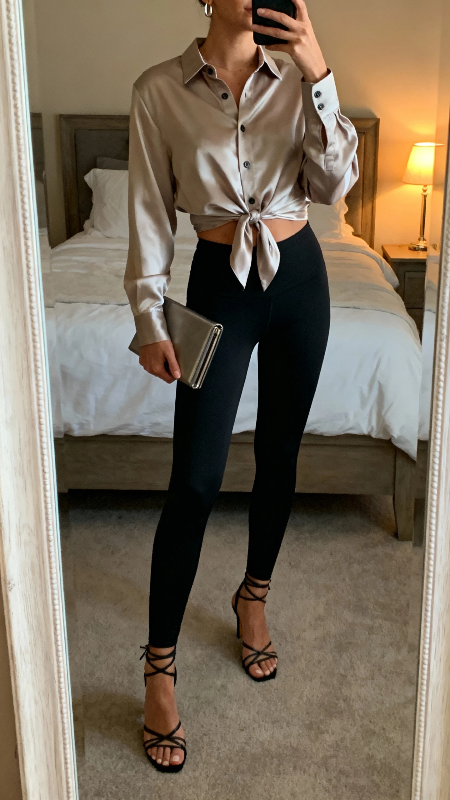 iPhone mirror selfie of a woman wearing a satin button-up shirt half-tucked into matte black leggings, strappy heeled sandals, slim clutch, subtle earrings, face not visible, cropped at neck, bedroom full-length mirror, warm evening lamp light, iPhone photo quality.