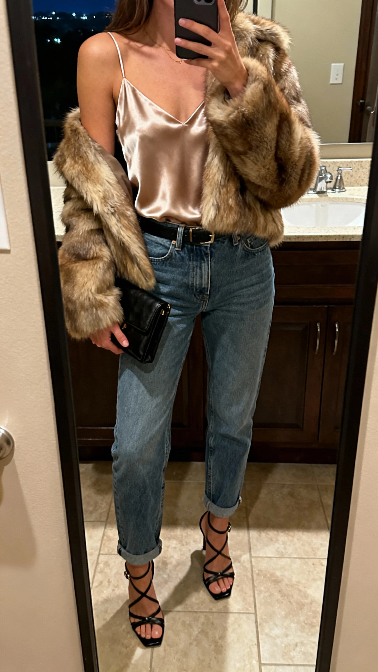 iPhone mirror selfie of a woman wearing a satin camisole tucked into mom jeans with a faux-fur jacket, strappy heels, and a clutch, face not visible, cropped at shoulders, bathroom mirror, evening indoor lighting, iPhone photo quality.