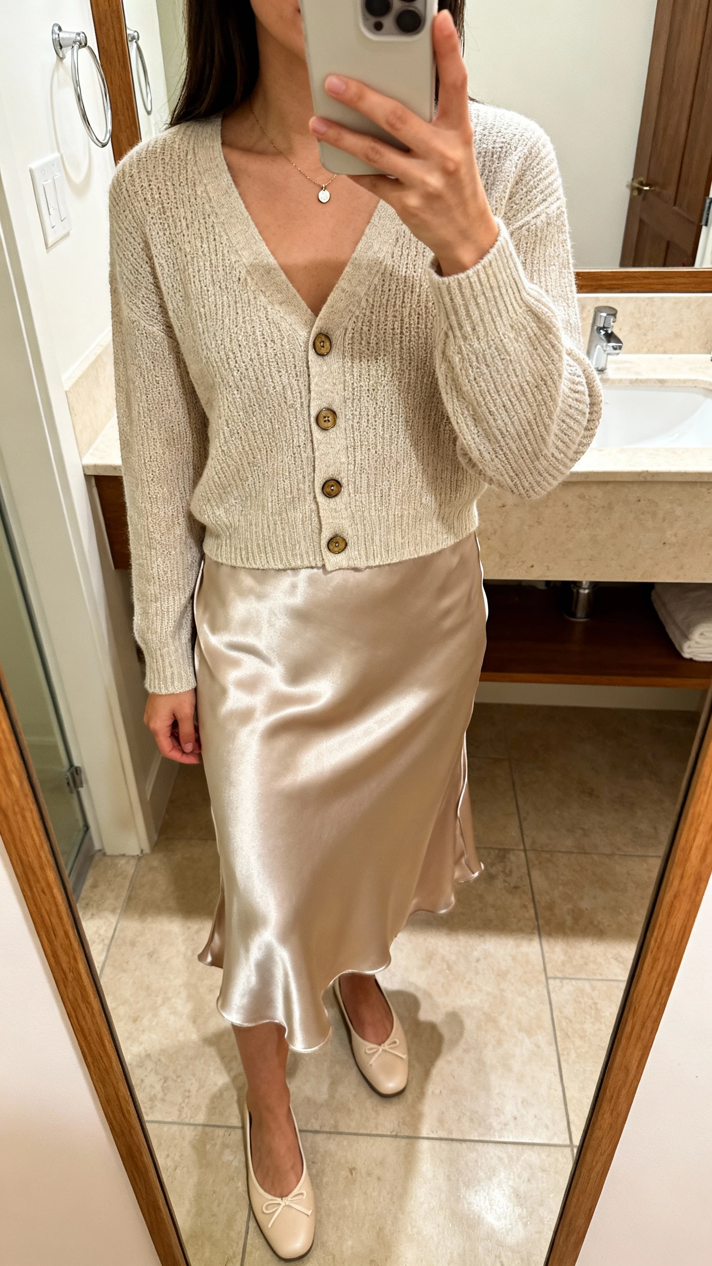 iPhone mirror selfie of a woman wearing a satin slip skirt with a matching fine-knit cardigan set (cardi worn buttoned), ballet flats, and a petite pendant necklace, face not visible, bathroom mirror, warm indoor lighting, iPhone photo quality.