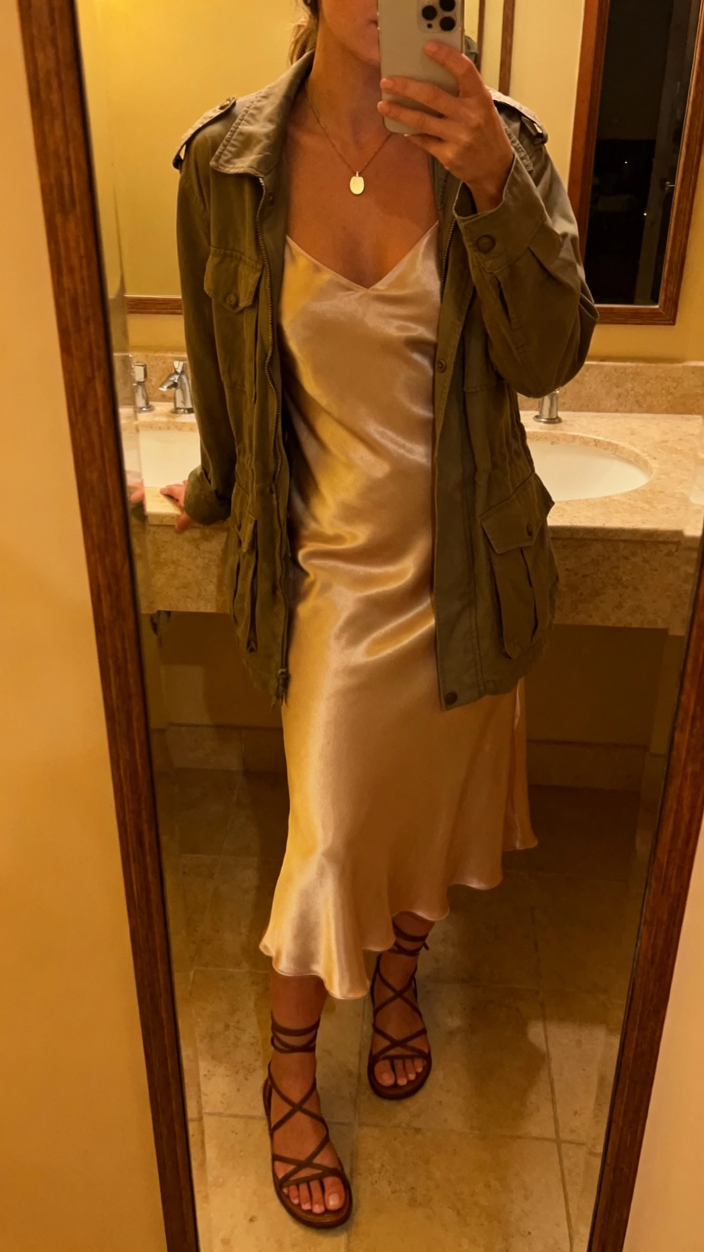 iPhone mirror selfie of a woman wearing a satin slip dress layered with a utility jacket, strappy sandals, and a simple pendant necklace, face not visible, bathroom mirror, evening indoor lighting, iPhone photo quality.