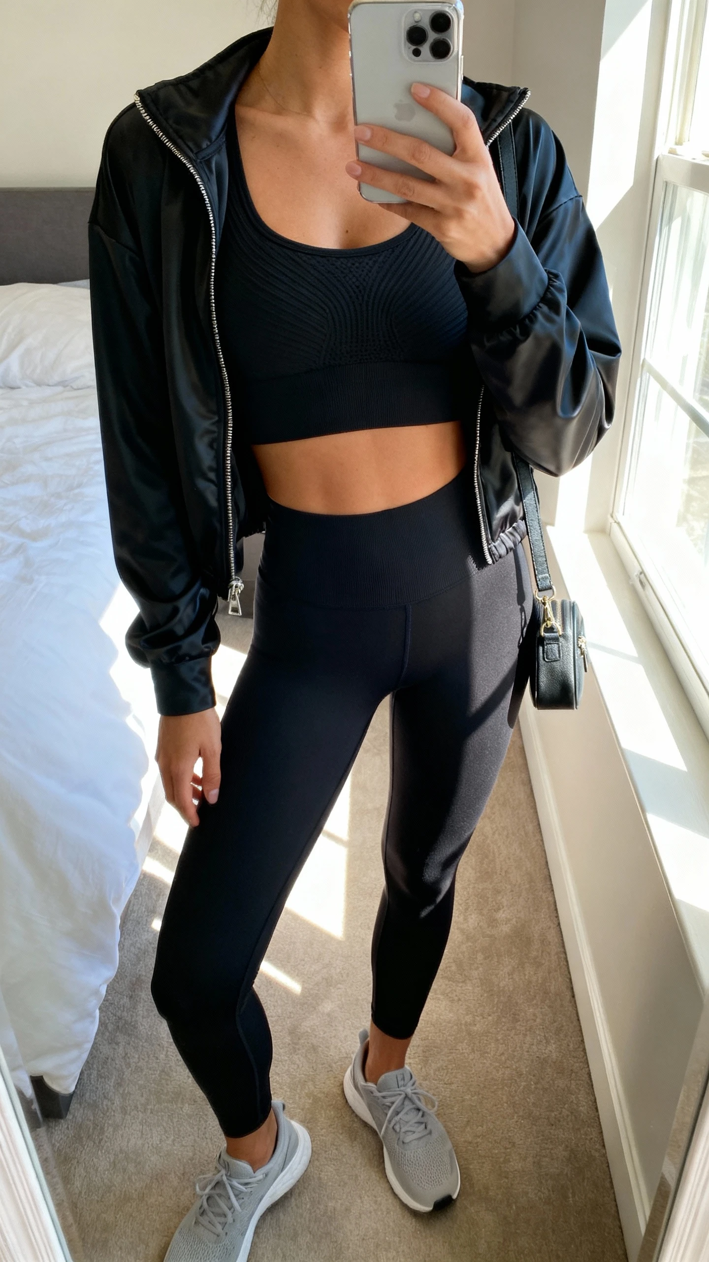 iPhone mirror selfie of a woman wearing a sculpted black seamless sports bra and matching high-waist leggings with a sleek black zip-up track jacket layered on top, face not visible, cropped at neck, bedroom mirror with natural window light, minimalist trainers and small crossbody, iPhone photo quality.