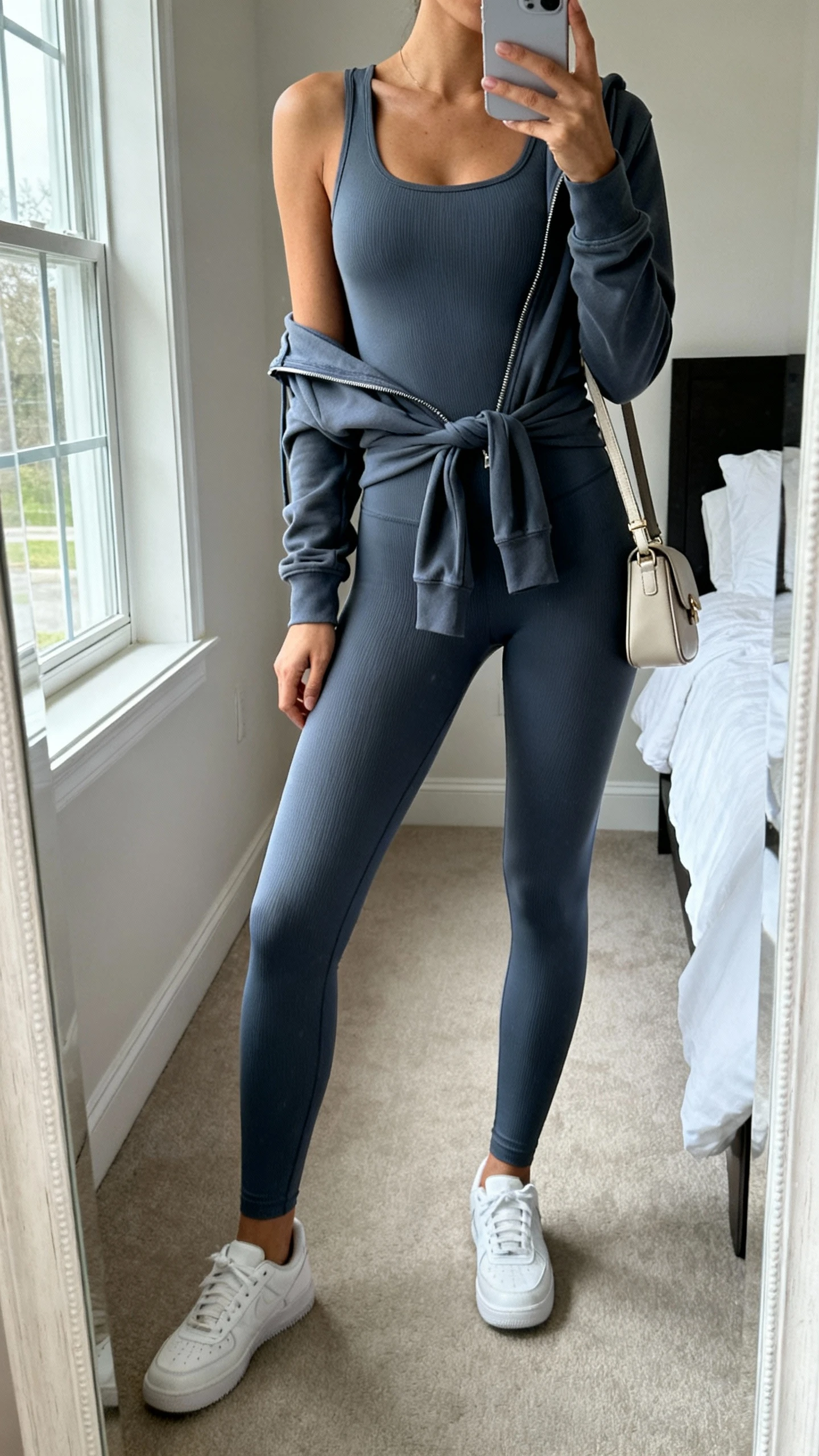 iPhone mirror selfie of a woman wearing a sculpted monochrome legging set (matching fitted leggings and longline tank), sleek zip-up hoodie tied at waist, minimal white sneakers, small structured crossbody, face not visible, cropped at neck, bedroom mirror, natural window light, iPhone photo quality.