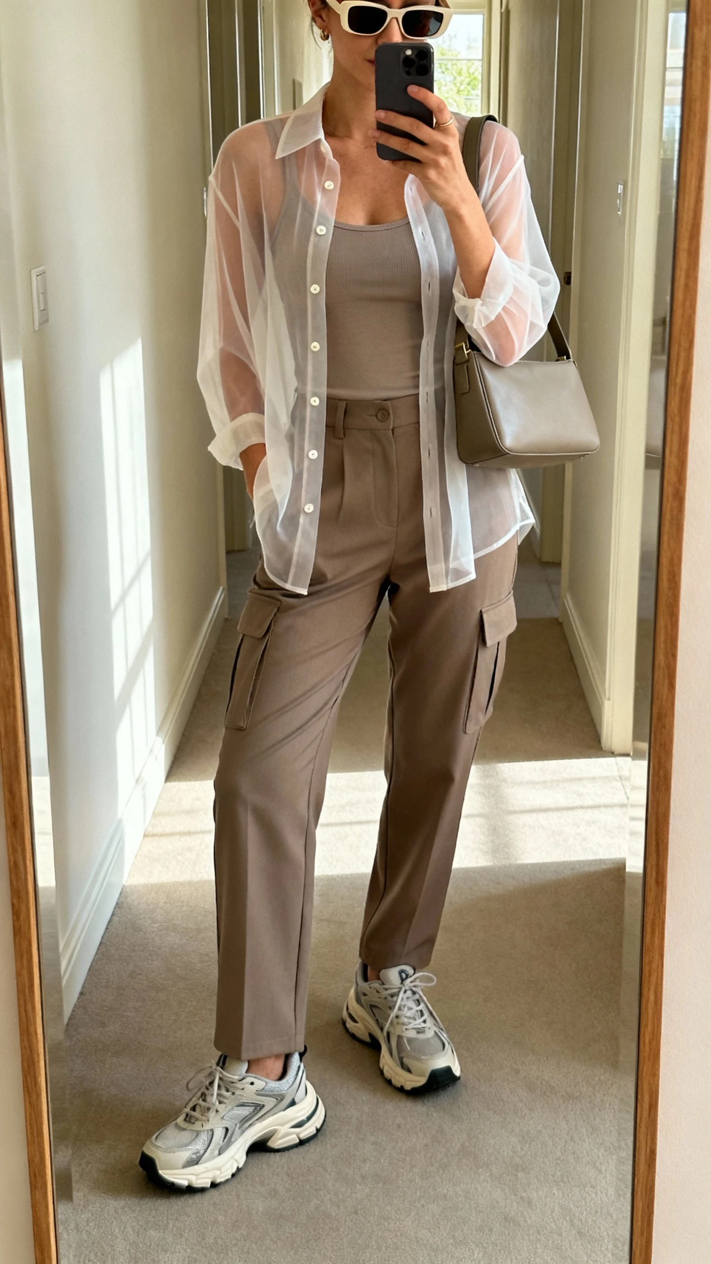 iPhone mirror selfie of a woman wearing a semi-sheer chiffon button-up blouse layered over a matching fitted tank, high-waist tailored cargo pants, chunky dad sneakers, rectangular sunglasses, and a medium shoulder bag with clean lines, face not visible, cropped at shoulders, hallway mirror with natural daylight, iPhone photo quality.