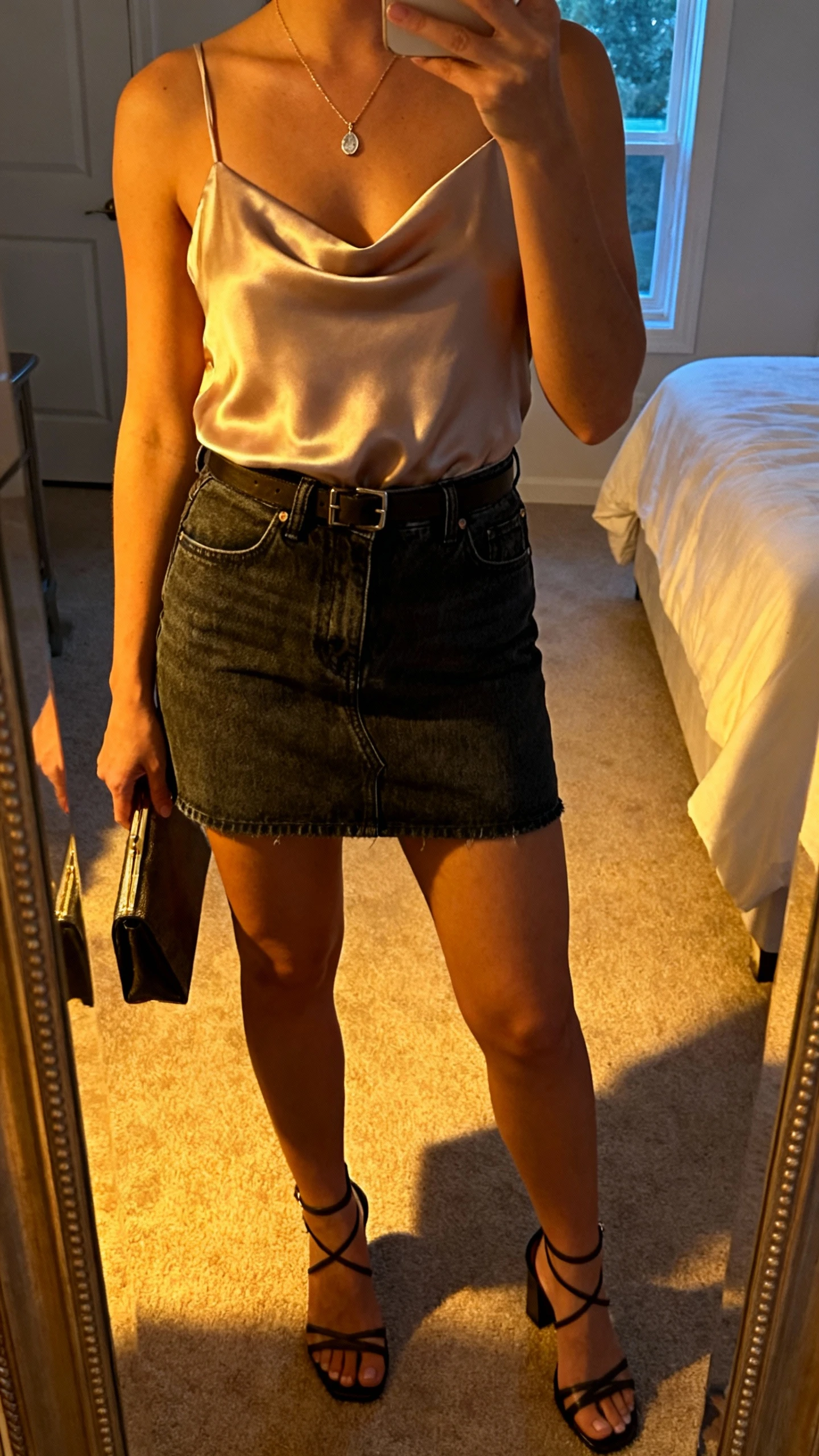 iPhone mirror selfie of a woman wearing a silk camisole tucked into a dark-wash denim mini skirt with strappy heeled sandals, delicate pendant necklace, and a small clutch, face not visible, bedroom mirror with warm evening light, iPhone photo quality.