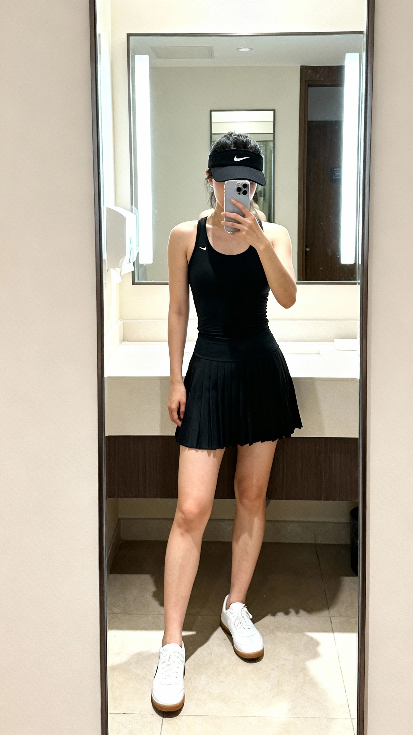 iPhone mirror selfie of a woman wearing a sleek black tennis skirt and a fitted black tank, face not visible, bathroom mirror with bright indoor lighting, black visor and white low-profile court sneakers, iPhone photo quality