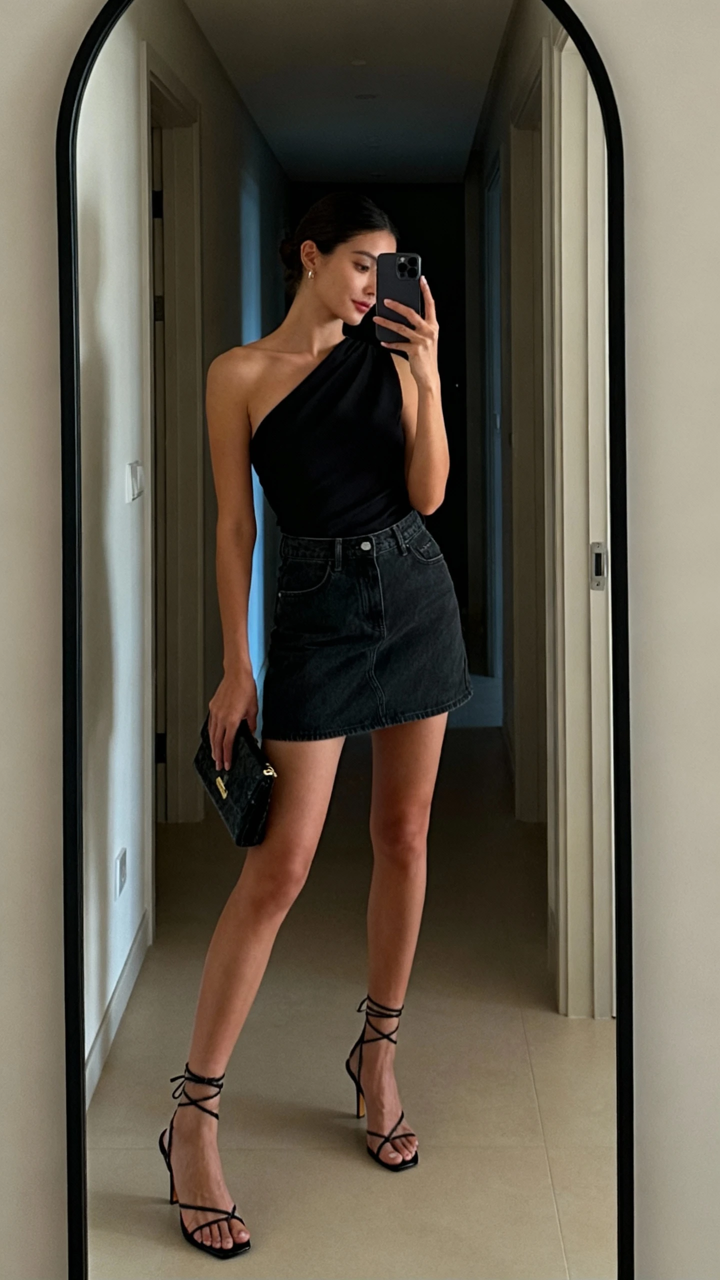 iPhone mirror selfie of a woman wearing a sleek one-shoulder black top with a dark denim mini skirt, strappy heeled sandals, and a small clutch, face not visible, side angle, hallway full-length mirror, evening indoor lighting, iPhone photo quality.