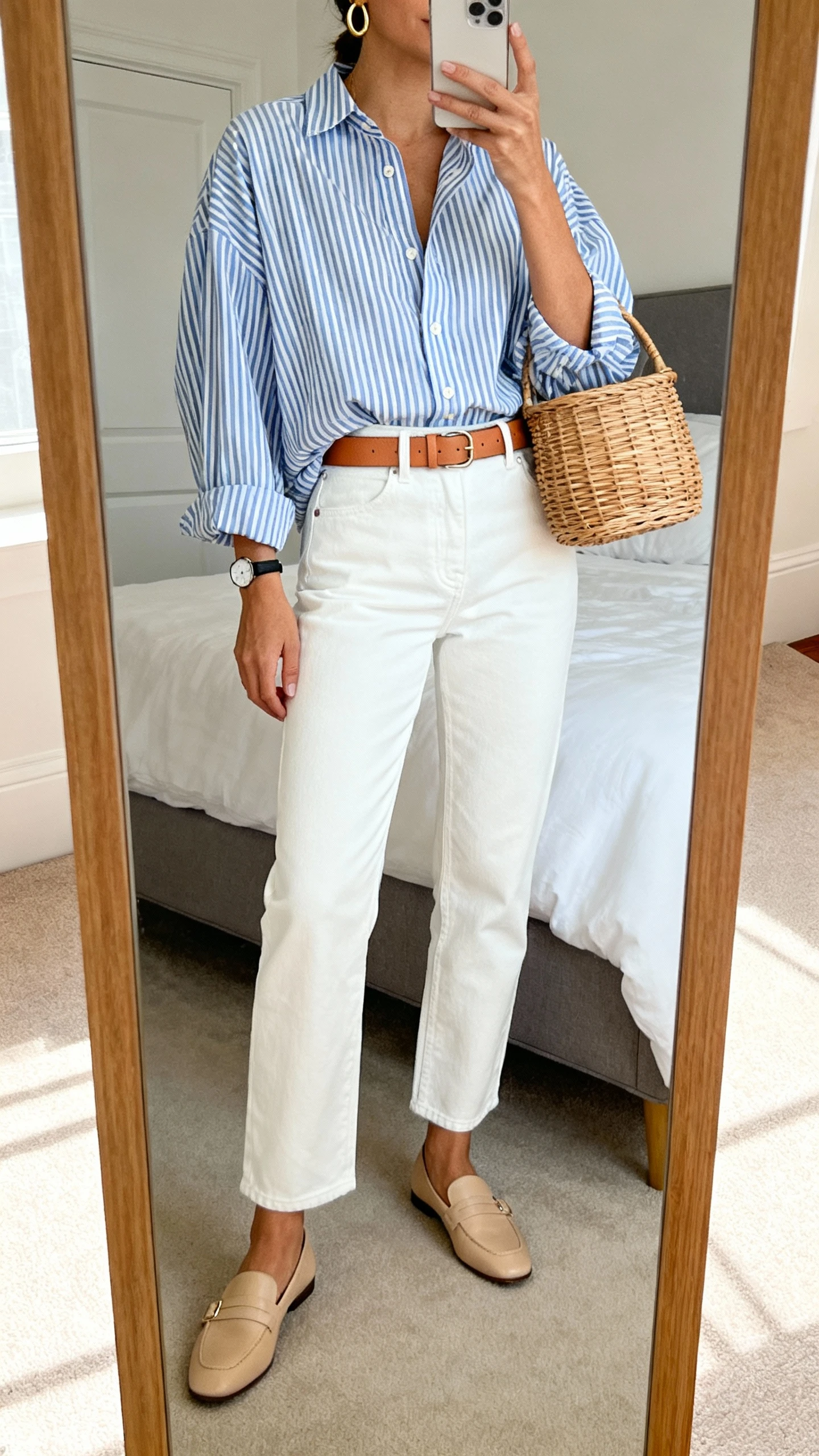 iPhone mirror selfie of a woman wearing a slightly oversized blue-and-white striped button-down, high-rise white straight-leg jeans, tan leather belt, beige loafers, structured basket bag, simple gold hoop earrings, and a slim watch, face not visible, cropped at neck, bedroom mirror with natural morning light, iPhone photo quality.