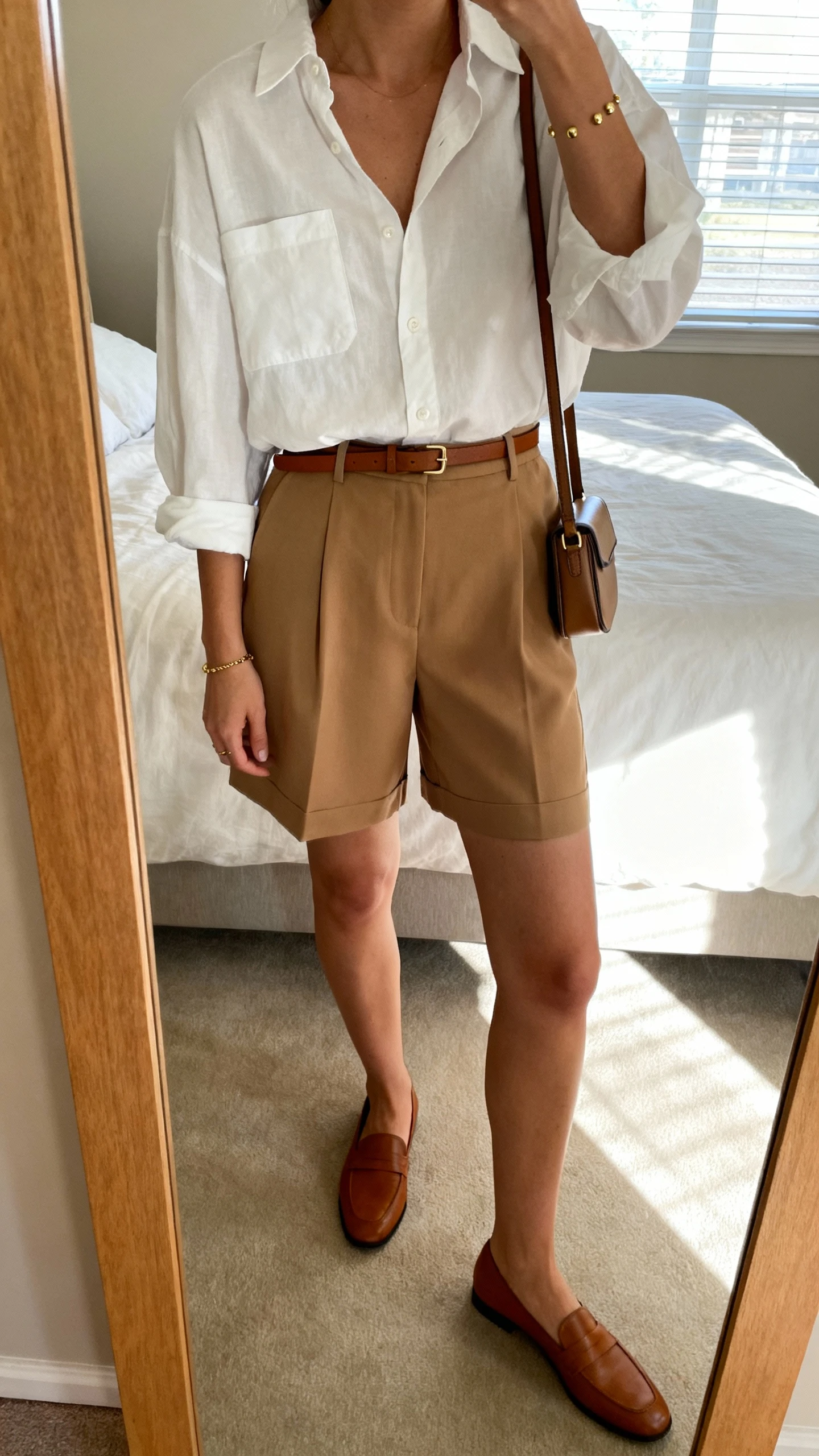 iPhone mirror selfie of a woman wearing a slightly oversized white cotton button-down tucked into high-waisted tan tailored shorts, thin brown belt, minimal leather loafers, structured mini crossbody, simple gold studs and bracelet, face not visible, cropped at neck, bedroom mirror, morning window light, iPhone photo quality.