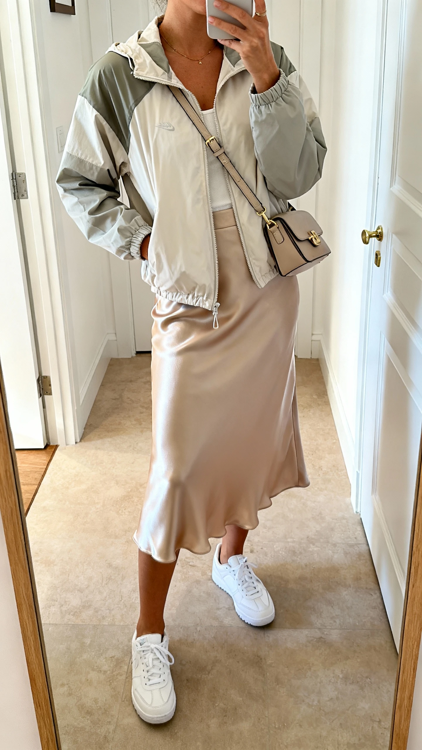 iPhone mirror selfie of a woman wearing a sporty-feminine mix: lightweight windbreaker over a satin midi skirt with sleek white sneakers and a crossbody bag, face not visible, cropped at neck, entryway mirror, bright indoor lighting, iPhone photo quality.