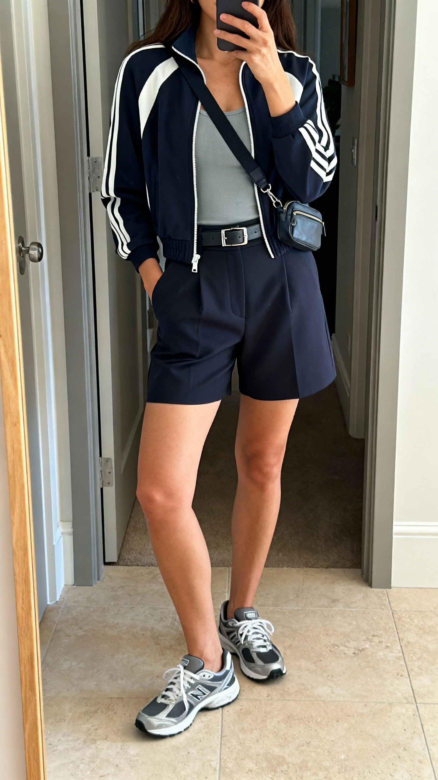 iPhone mirror selfie of a woman wearing a sporty track jacket zipped halfway over a fitted tank, tailored high-waist shorts, sleek trainers, and a crossbody belt bag, face not visible, entryway mirror, natural daylight, iPhone photo quality.