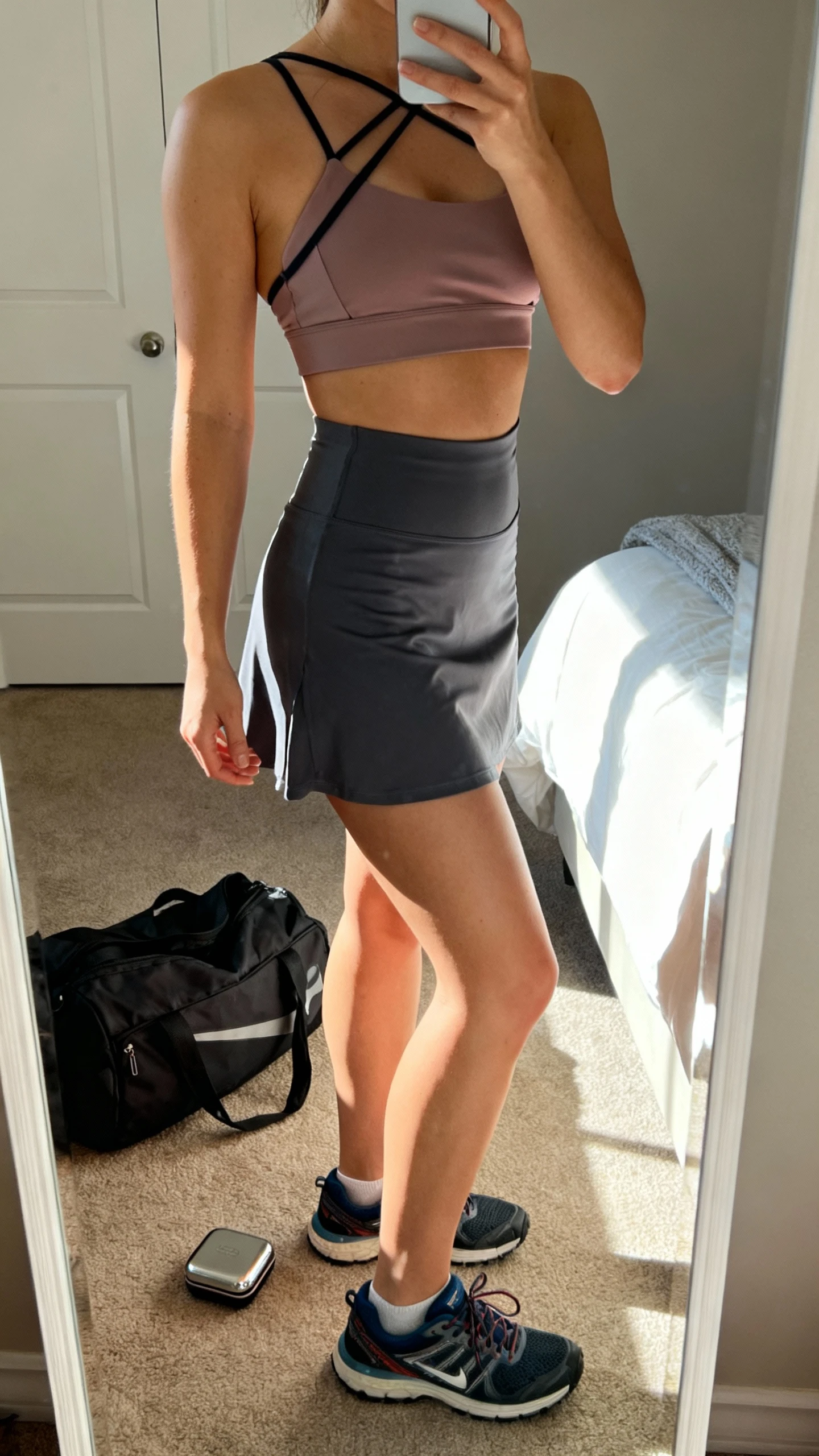 iPhone mirror selfie of a woman wearing a strappy sports bra and high-rise training skort, face not visible, bedroom mirror with morning light, cross-training sneakers and a compact gym bag on the floor, iPhone photo quality.
