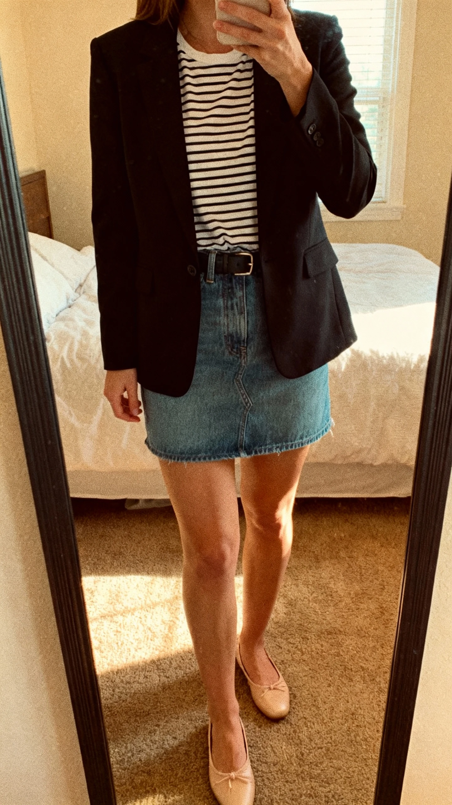 iPhone mirror selfie of a woman wearing a striped tee tucked into a denim mini skirt, tailored black blazer, and ballet flats, face not visible, cropped at neck, bedroom mirror, soft morning window light, iPhone photo quality.