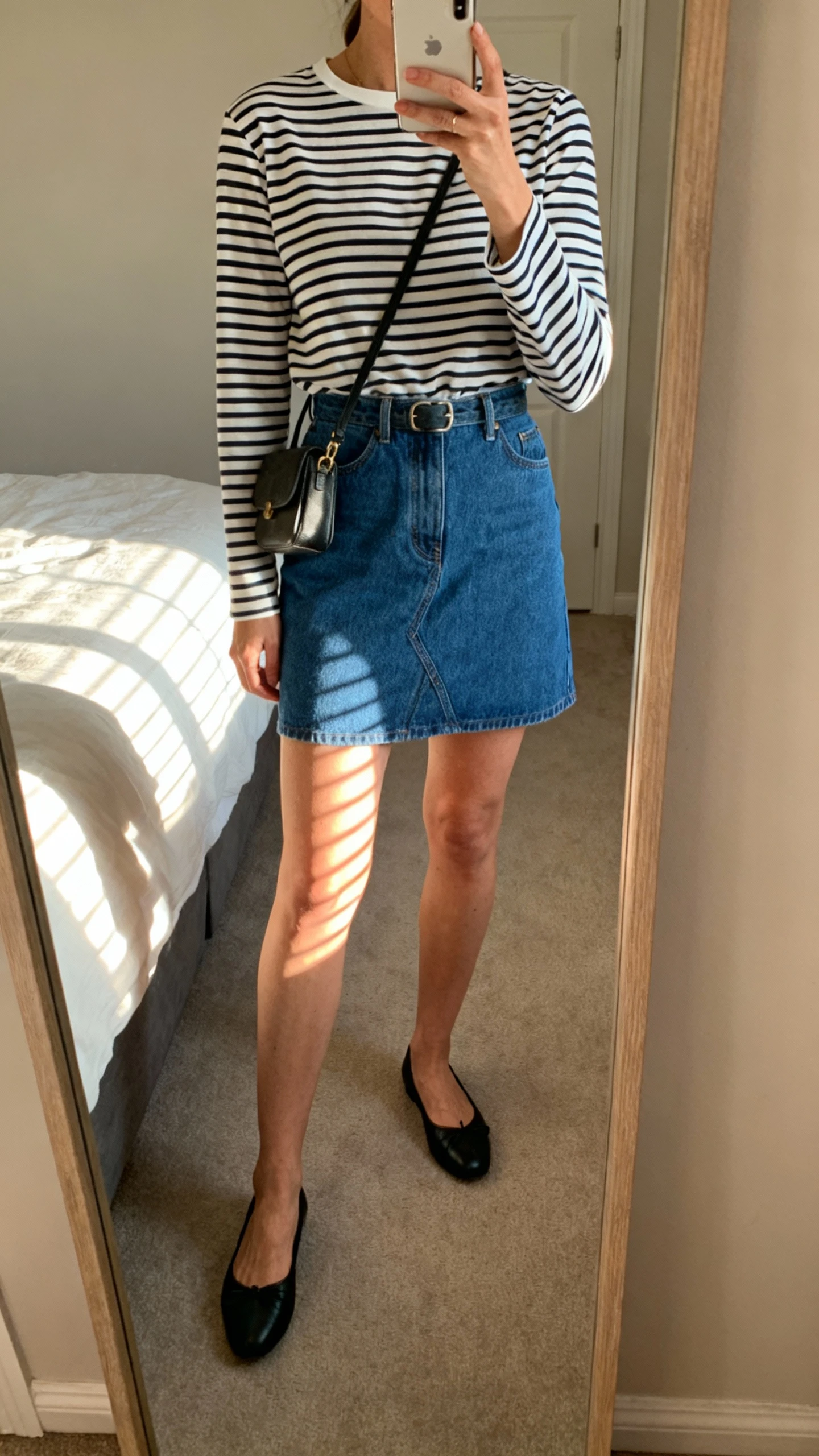 iPhone mirror selfie of a woman wearing a striped Breton long-sleeve tee tucked into a blue denim mini skirt with black ballet flats and a small crossbody bag, face not visible, cropped at neck, bedroom mirror with morning window light, iPhone photo quality.