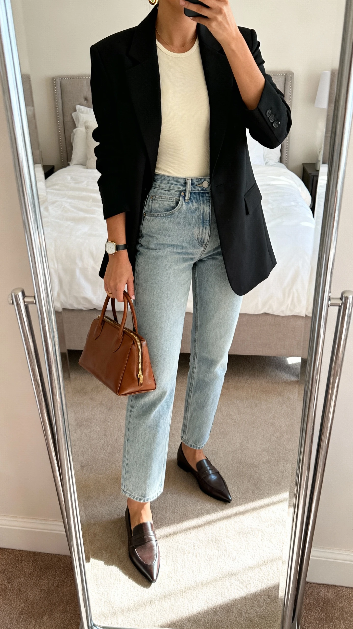 iPhone mirror selfie of a woman wearing a tailored black blazer, high-rise straight-leg light-wash jeans, a fitted cream bodysuit, pointed-toe loafers, minimal gold hoops, slim watch, and a structured leather tote in hand; face not visible, cropped at neck, bedroom mirror with natural morning light, iPhone photo quality.