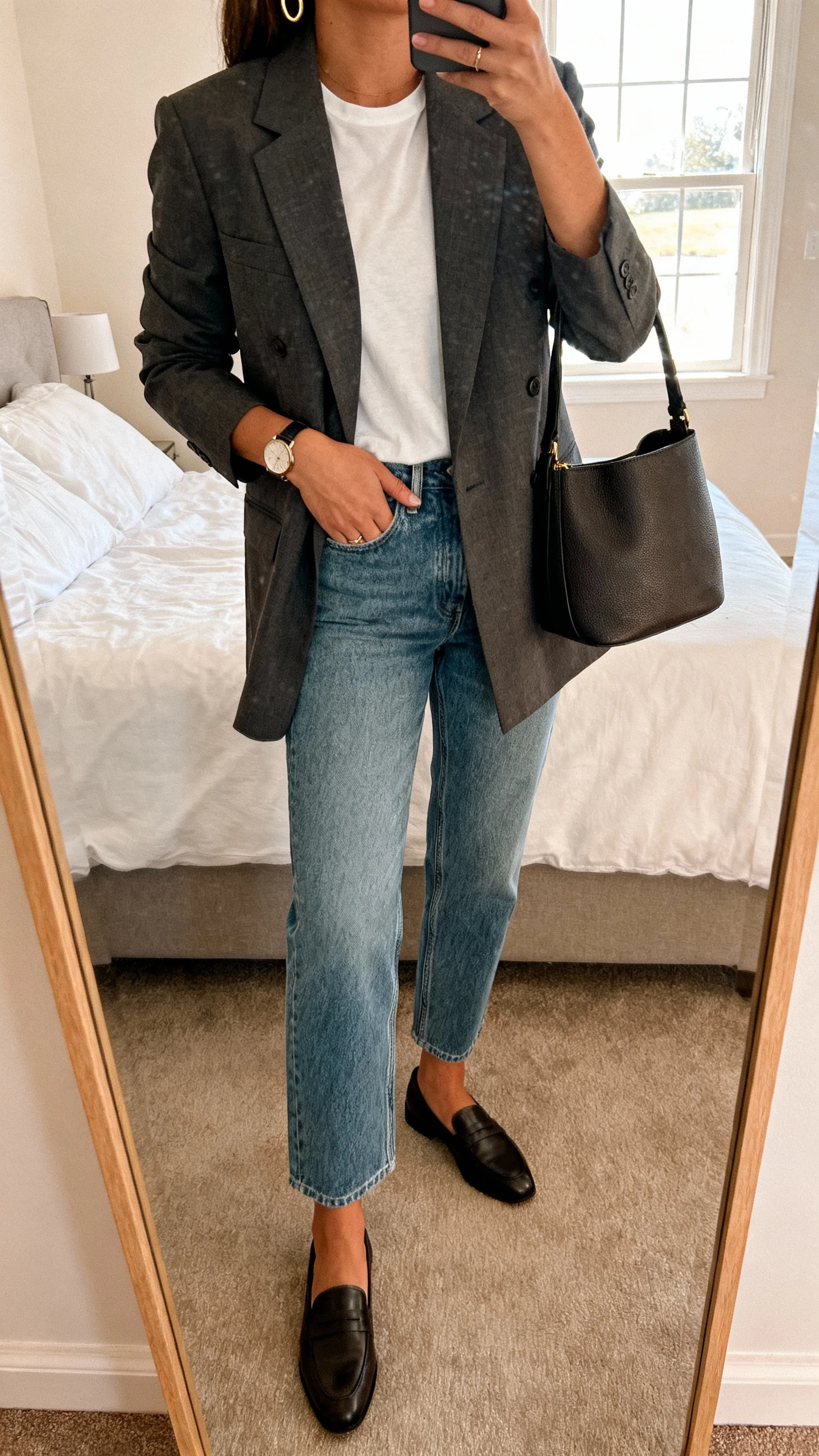 iPhone mirror selfie of a woman wearing a tailored blazer over a simple tee, clean straight-leg denim, sleek loafers, structured tote, minimal gold hoops and a watch, face not visible, cropped at neck, bedroom mirror, morning window light, iPhone photo quality.