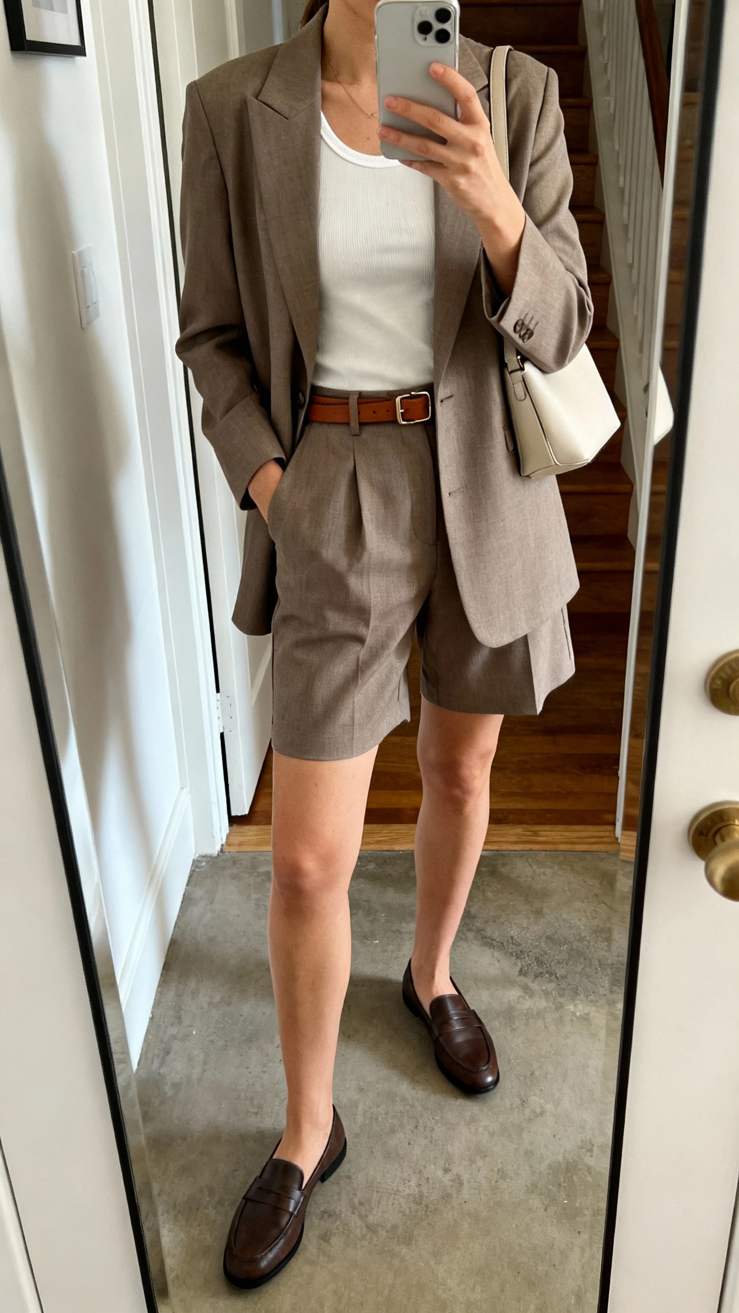 iPhone mirror selfie of a woman wearing a tailored shorts suit (structured blazer and matching tailored shorts) with a fitted tank, loafers, slim leather belt, minimalist shoulder bag, face not visible, cropped at neck, apartment entry mirror, natural daylight, iPhone photo quality.