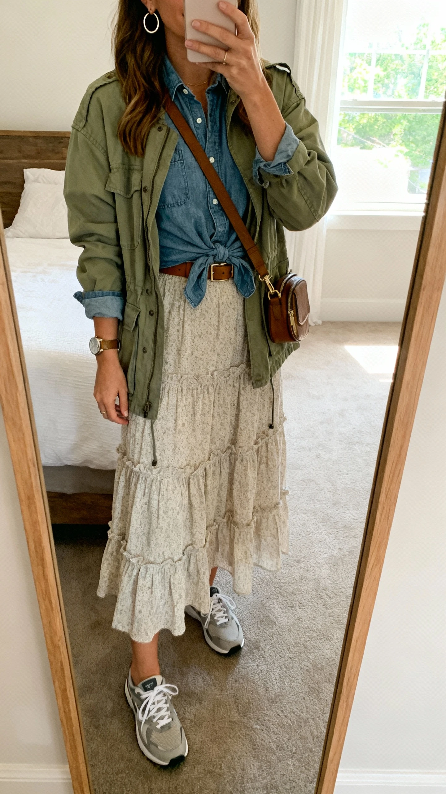 iPhone mirror selfie of a woman wearing a tiered midi dress with a chambray shirt tied at the waist and a lightweight olive utility jacket, low-profile sneakers, a crossbody belt bag, simple hoop earrings and a watch, face not visible, bedroom mirror, afternoon window light, iPhone photo quality.