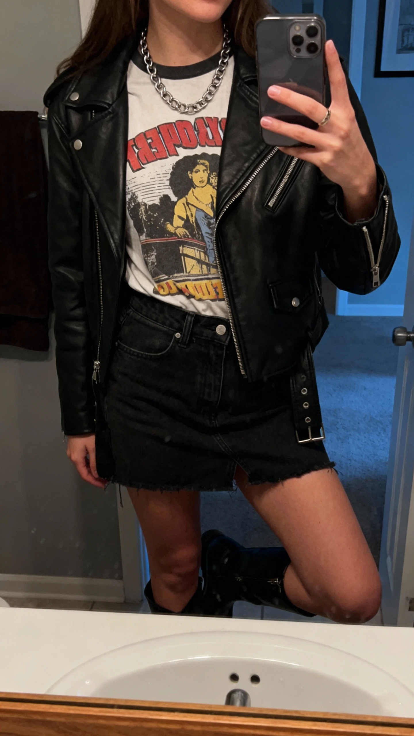 iPhone mirror selfie of a woman wearing a vintage graphic tee tucked into a black denim mini skirt with a black leather moto jacket, ankle boots, and chain necklace, face not visible, bathroom mirror, moody indoor lighting, iPhone photo quality.