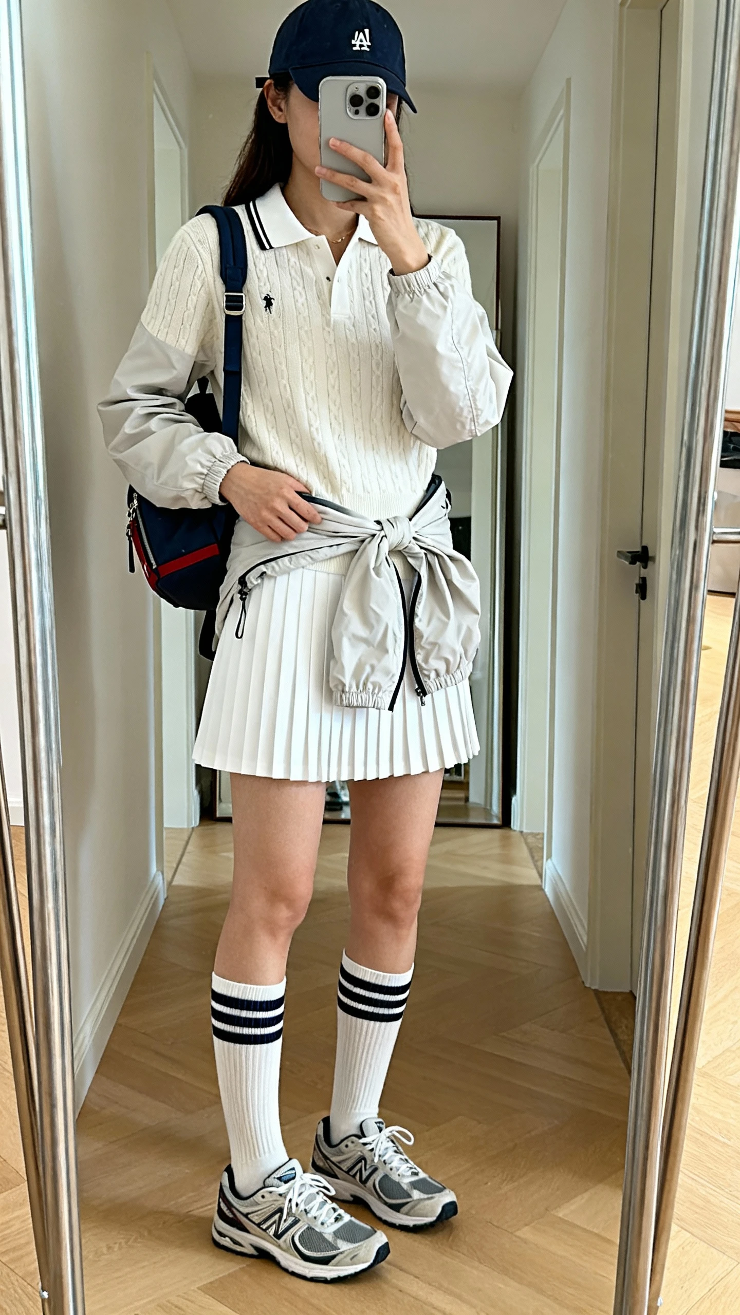 iPhone mirror selfie of a woman wearing a white pleated tennis skirt, collared knit polo, retro sneakers with mid-calf crew socks, mini backpack, baseball cap, and a lightweight windbreaker tied at the waist; face not visible, cropped at shoulders, full-length mirror in hallway, natural daylight, iPhone photo quality.